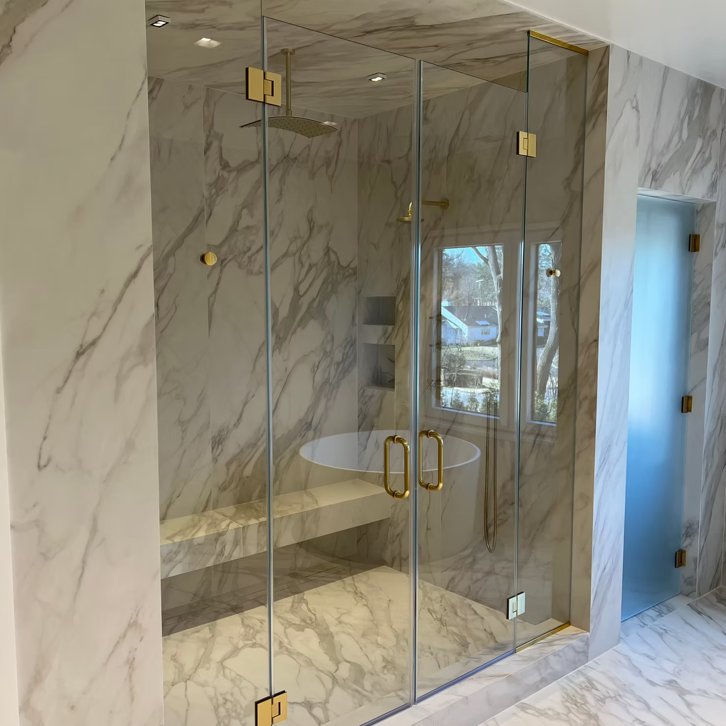 Glass shower enclosure with gold hinges in a marble-tiled bathroom