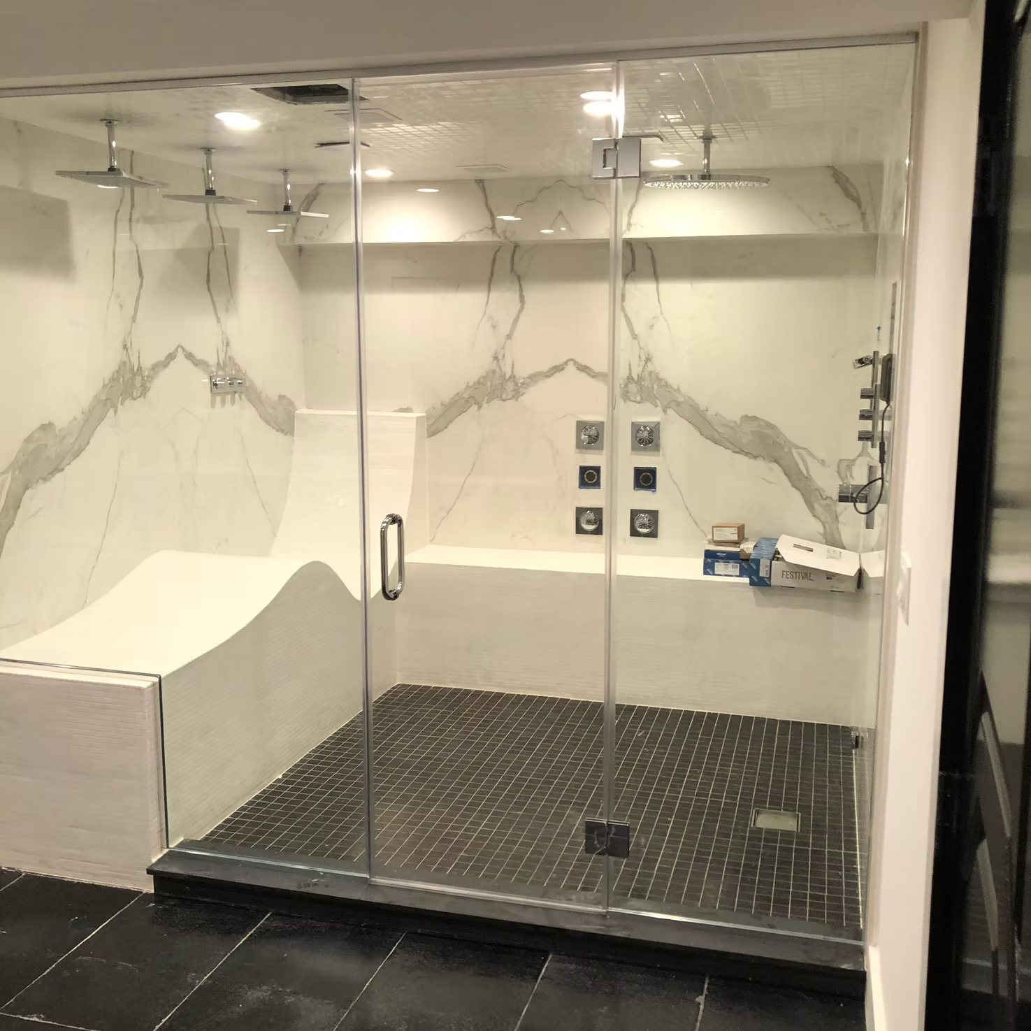 Glass-walled shower room with white fixtures, dark tile floor, and wall-mounted controls in a modern spa setting