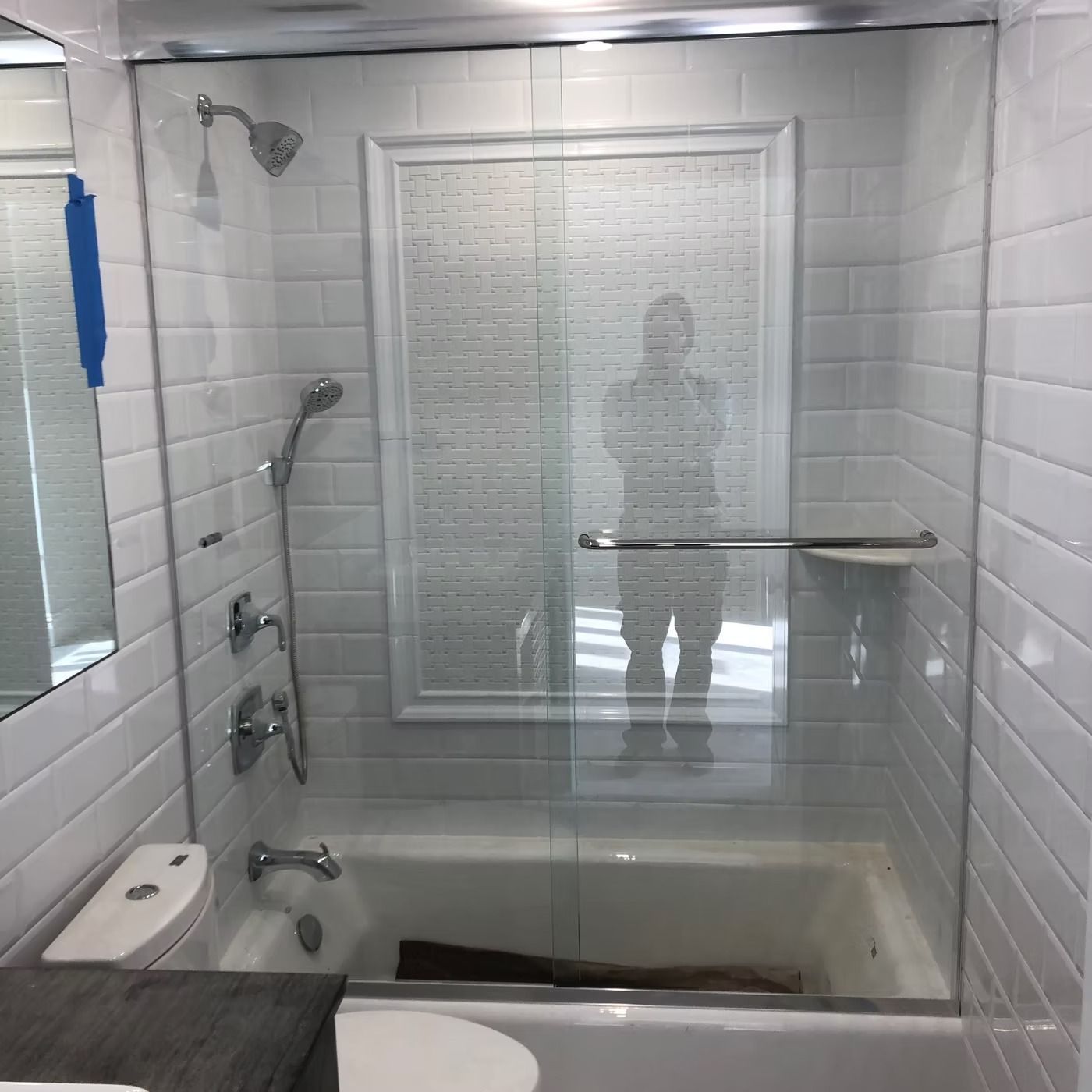 White-tiled bathroom with a glass shower, tub, and window reflected in the shower door.