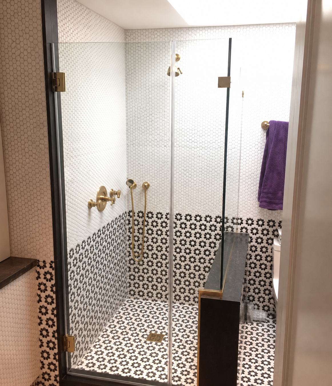 Walk-in shower with patterned tile walls, glass door, gold fixtures, and a purple towel.
