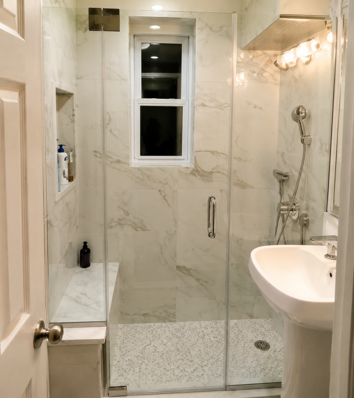 Small modern bathroom with glass shower, white tile, pedestal sink, and window.