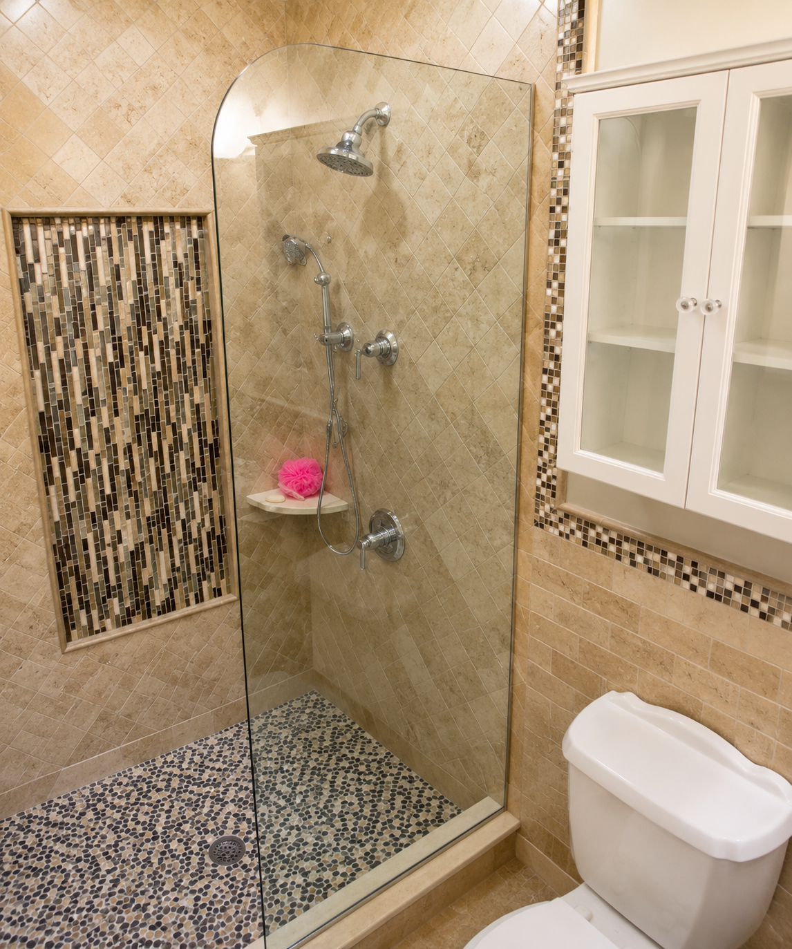 Walk-in tiled shower with glass panel, beige walls, mosaic floor, and a toilet beside a cabinet window.