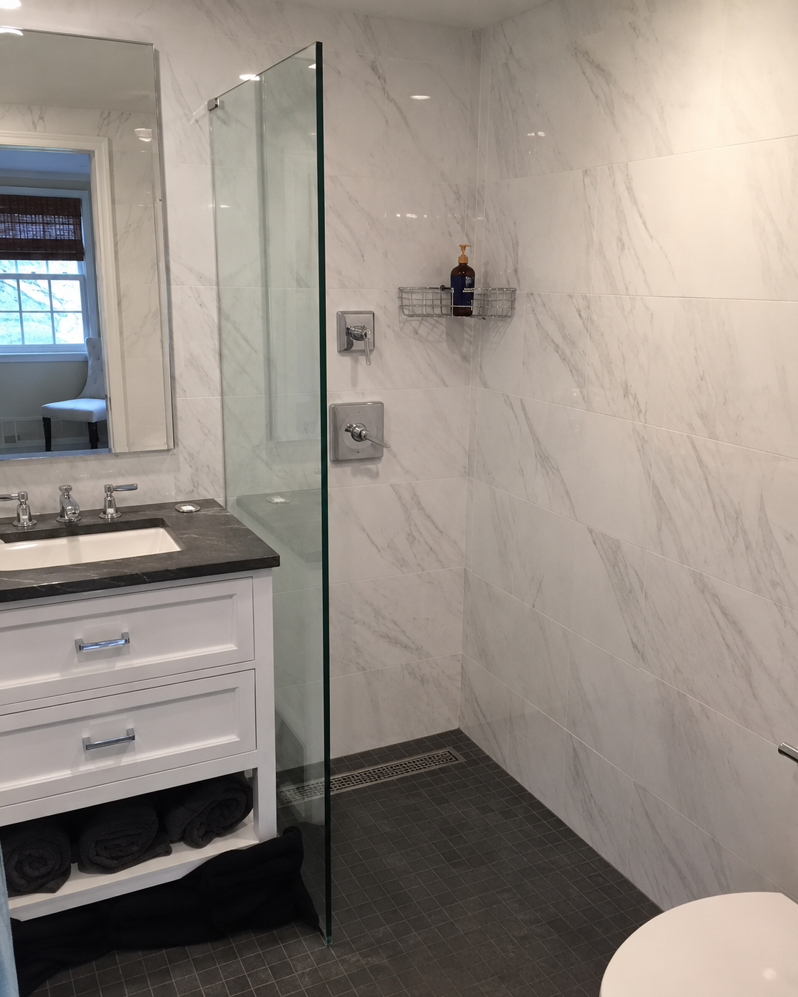 Modern bathroom with marble walls, dark tile floor, glass shower, vanity sink, and toilet edge visible.