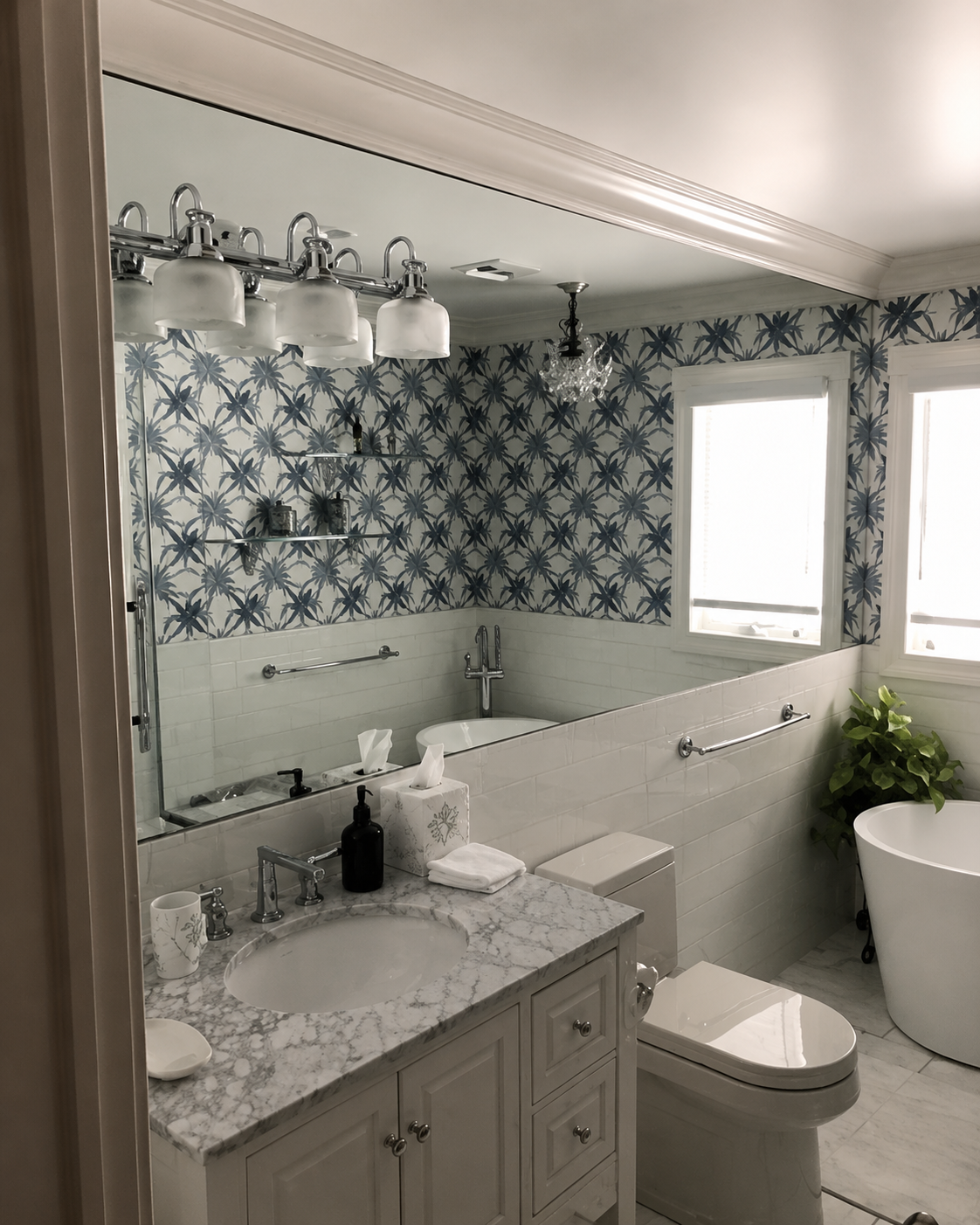 Bright bathroom with blue patterned wallpaper, freestanding tub, vanity, mirror, and potted plant by the window