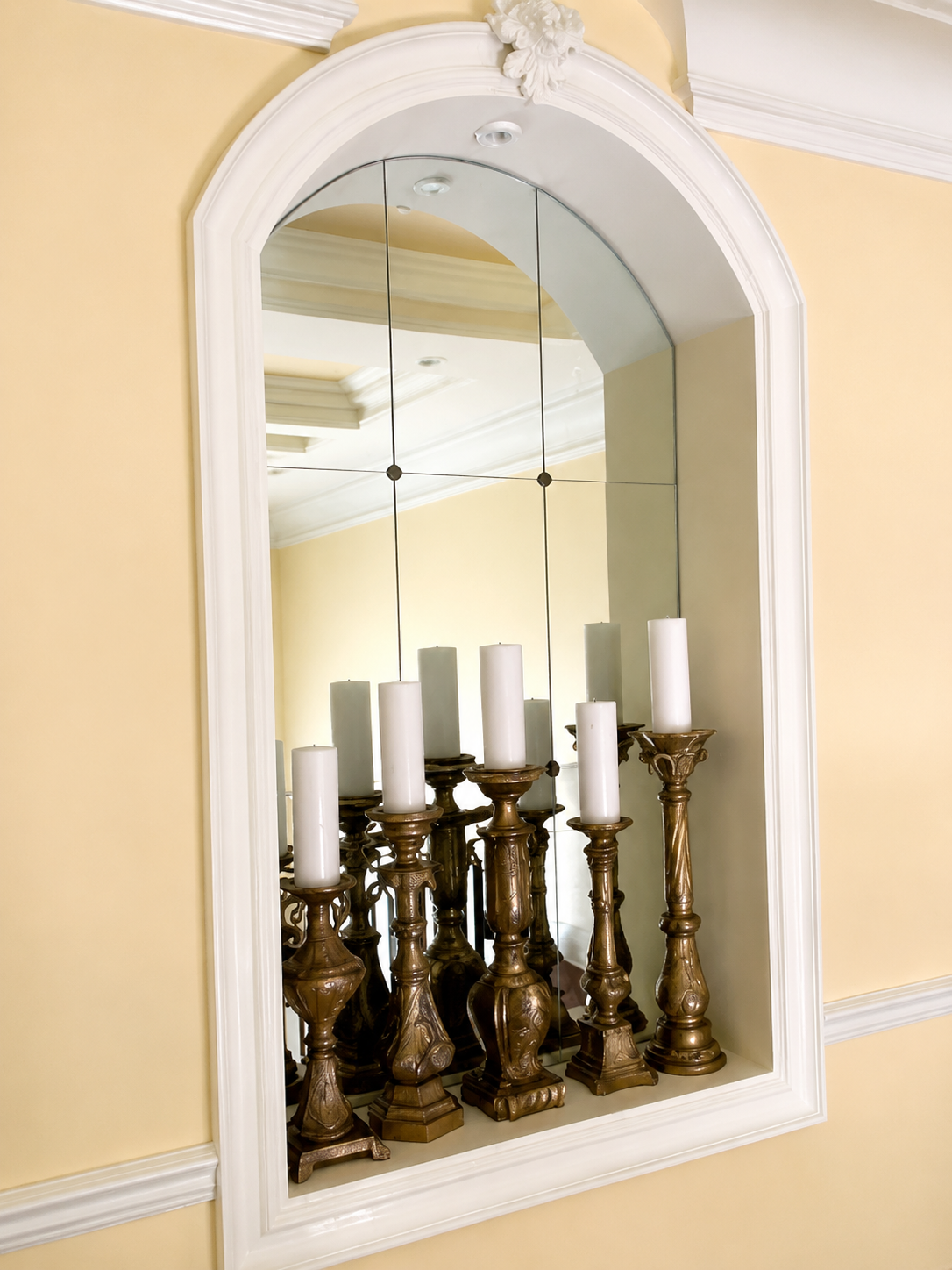 Decorative wall mirror reflecting silver candlesticks on a mantel, framed by a white arch on a beige wall