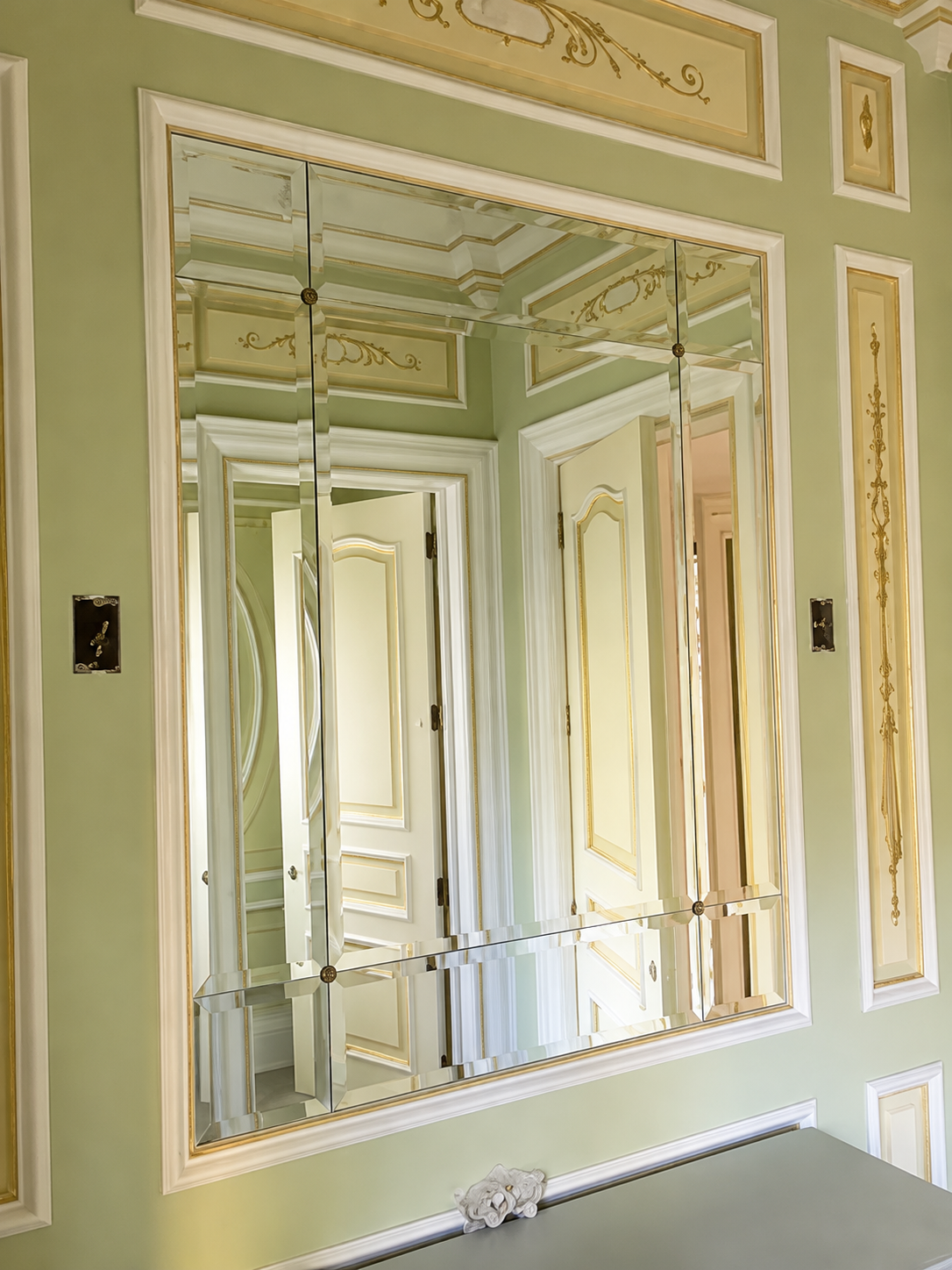 Ornate gold-trimmed wall mirror reflecting a bright, elegant room with paneled doors and pale green walls