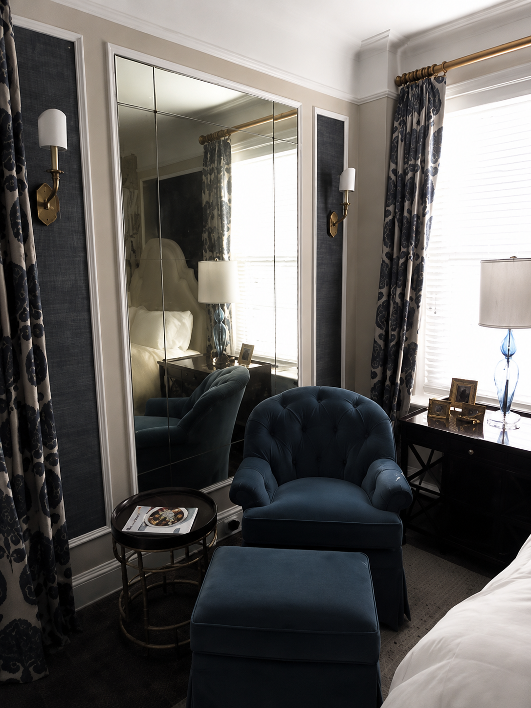 Elegant bedroom seating area with blue velvet chair, mirrored closet, window, and black side table.