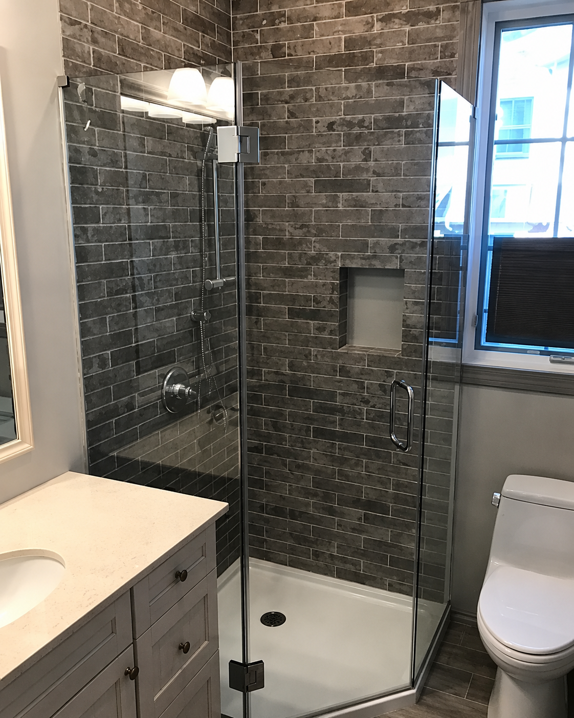 Modern bathroom with glass shower, white vanity, brick tile walls, and toilet beside a window.