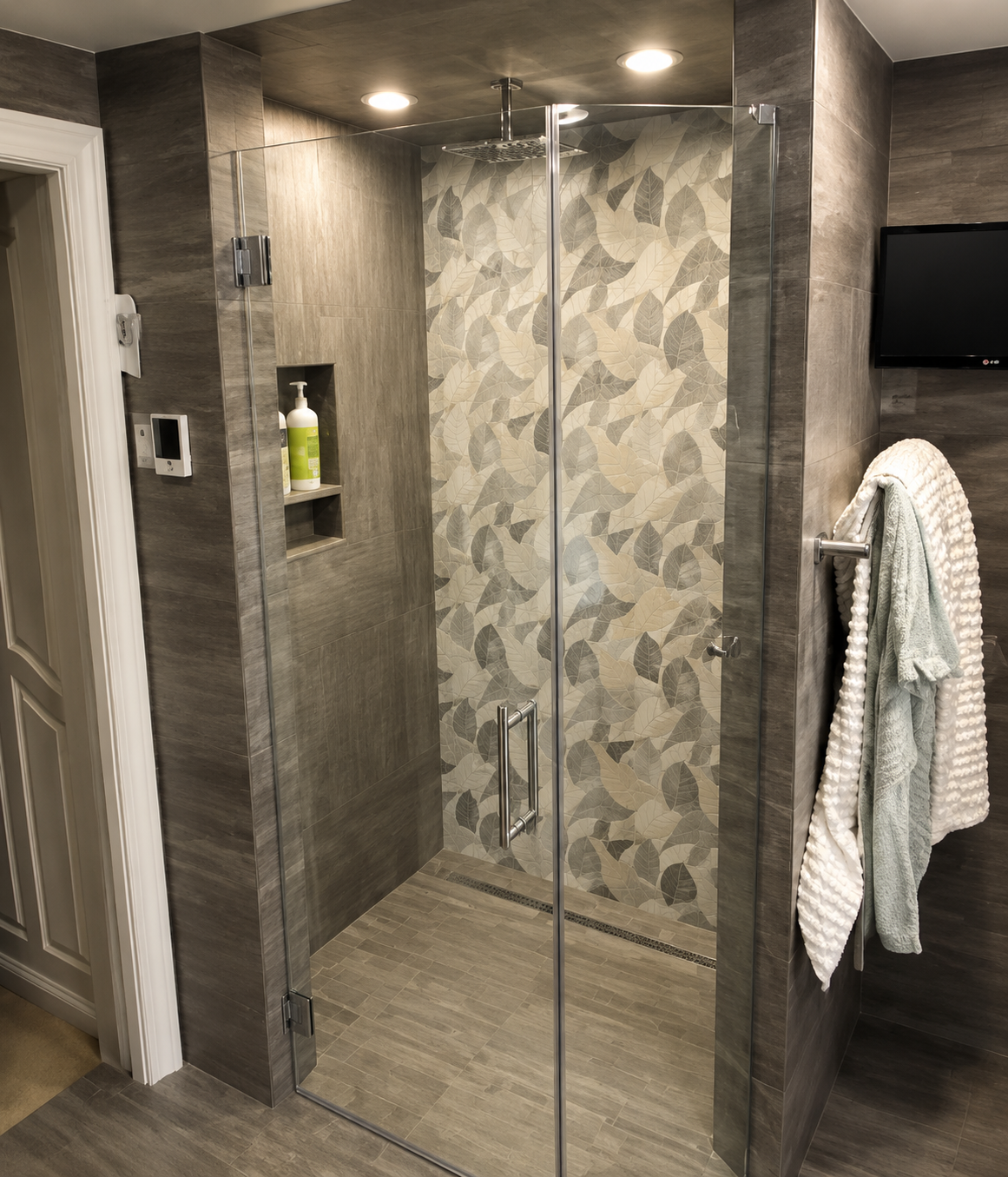 Modern glass shower with gray tile and patterned wall in a bathroom, with towels hanging nearby