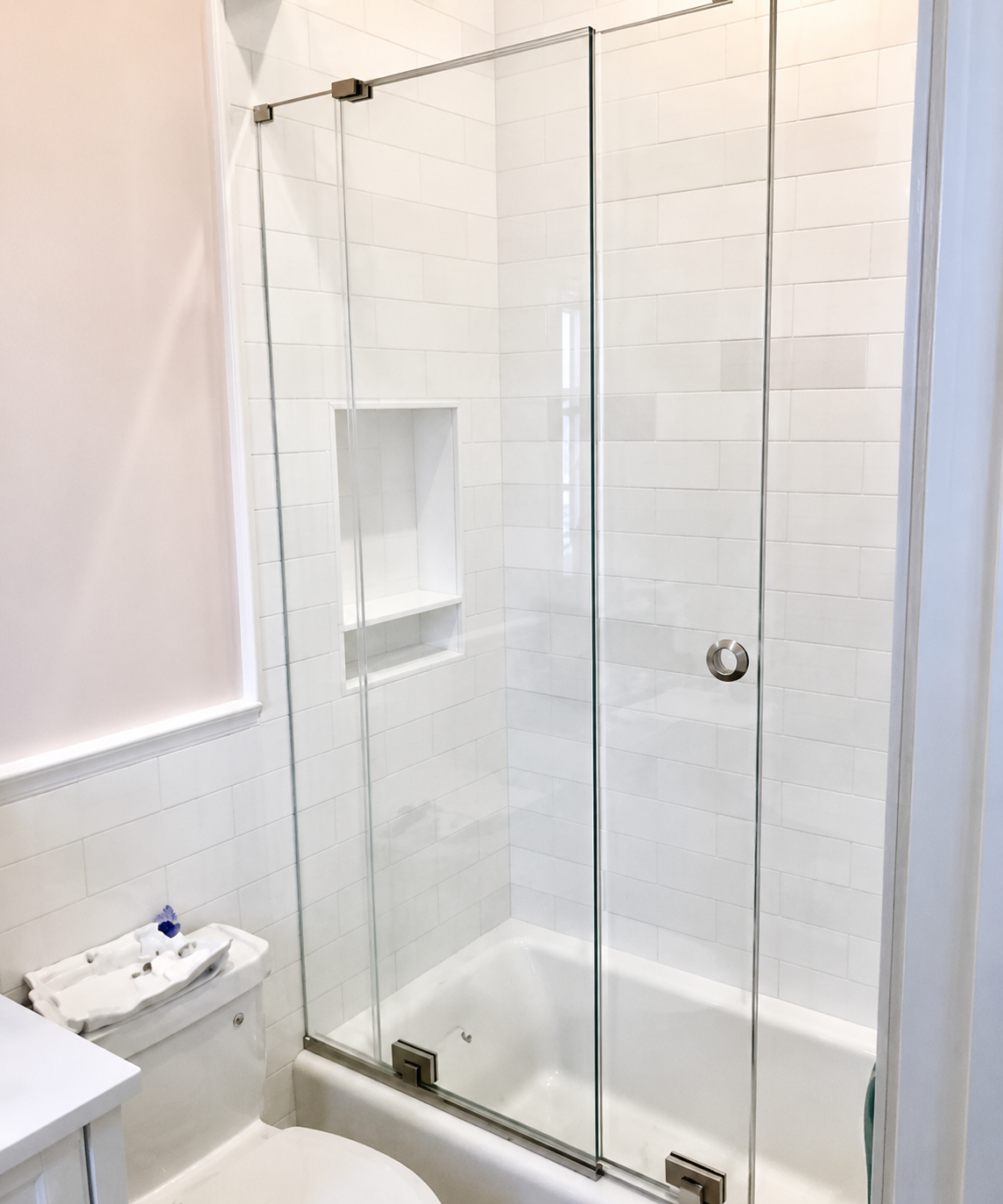 Glass-enclosed shower in a white tiled bathroom with a small sink and toilet nearby