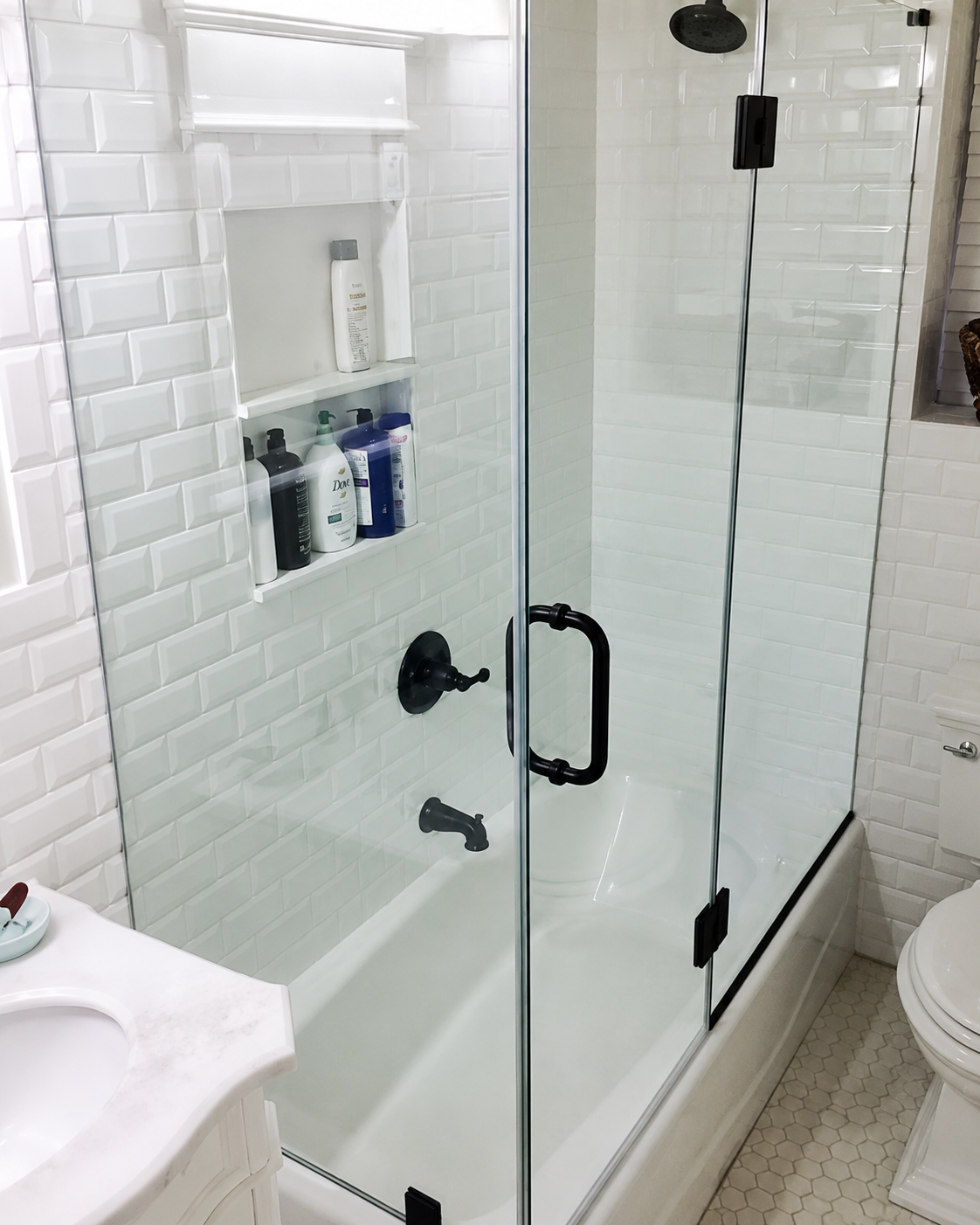 Glass shower enclosure with black hardware in a white-tiled bathroom, next to a toilet and sink