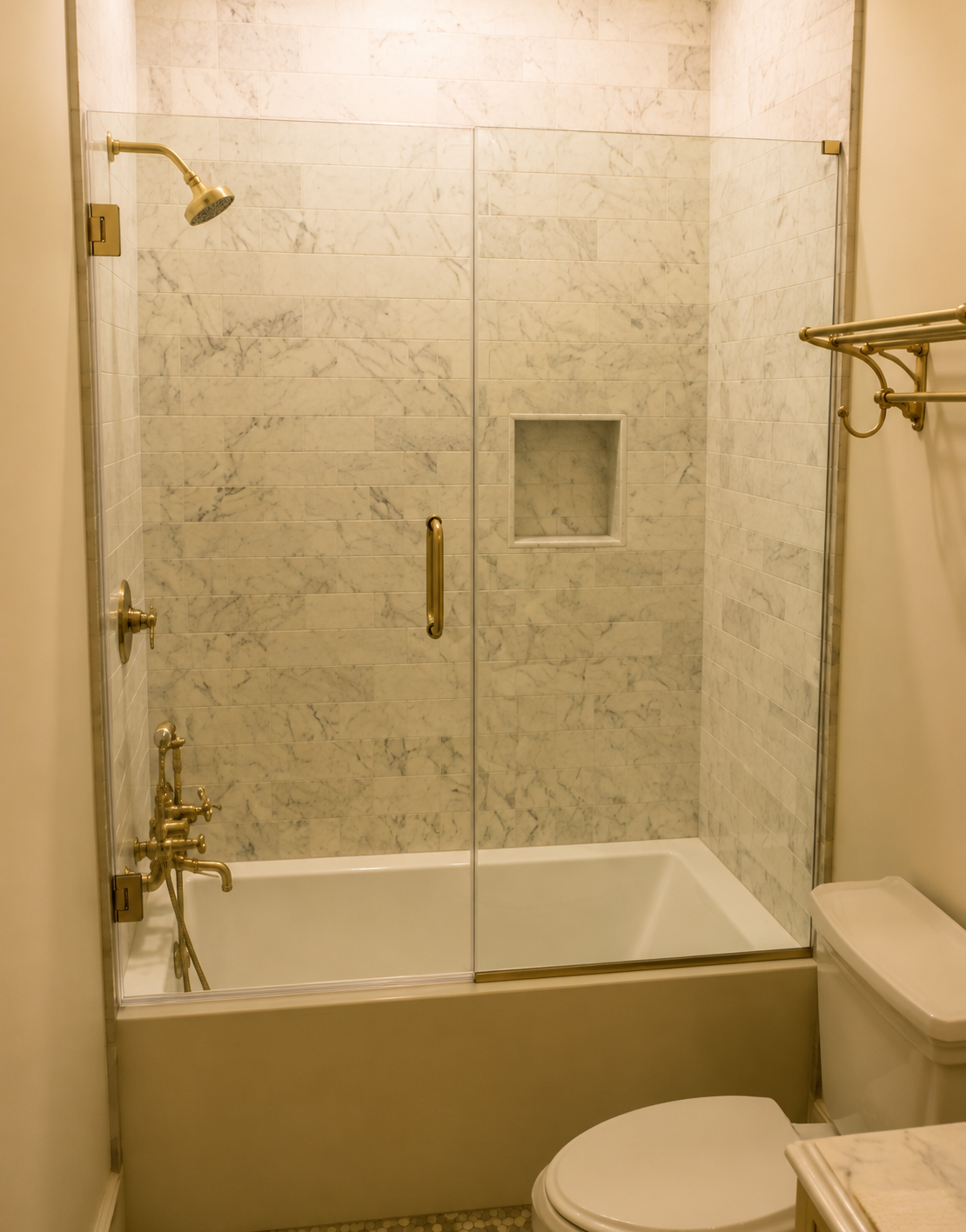 Small beige bathroom with a glass-enclosed bathtub shower, toilet, and brass fixtures.