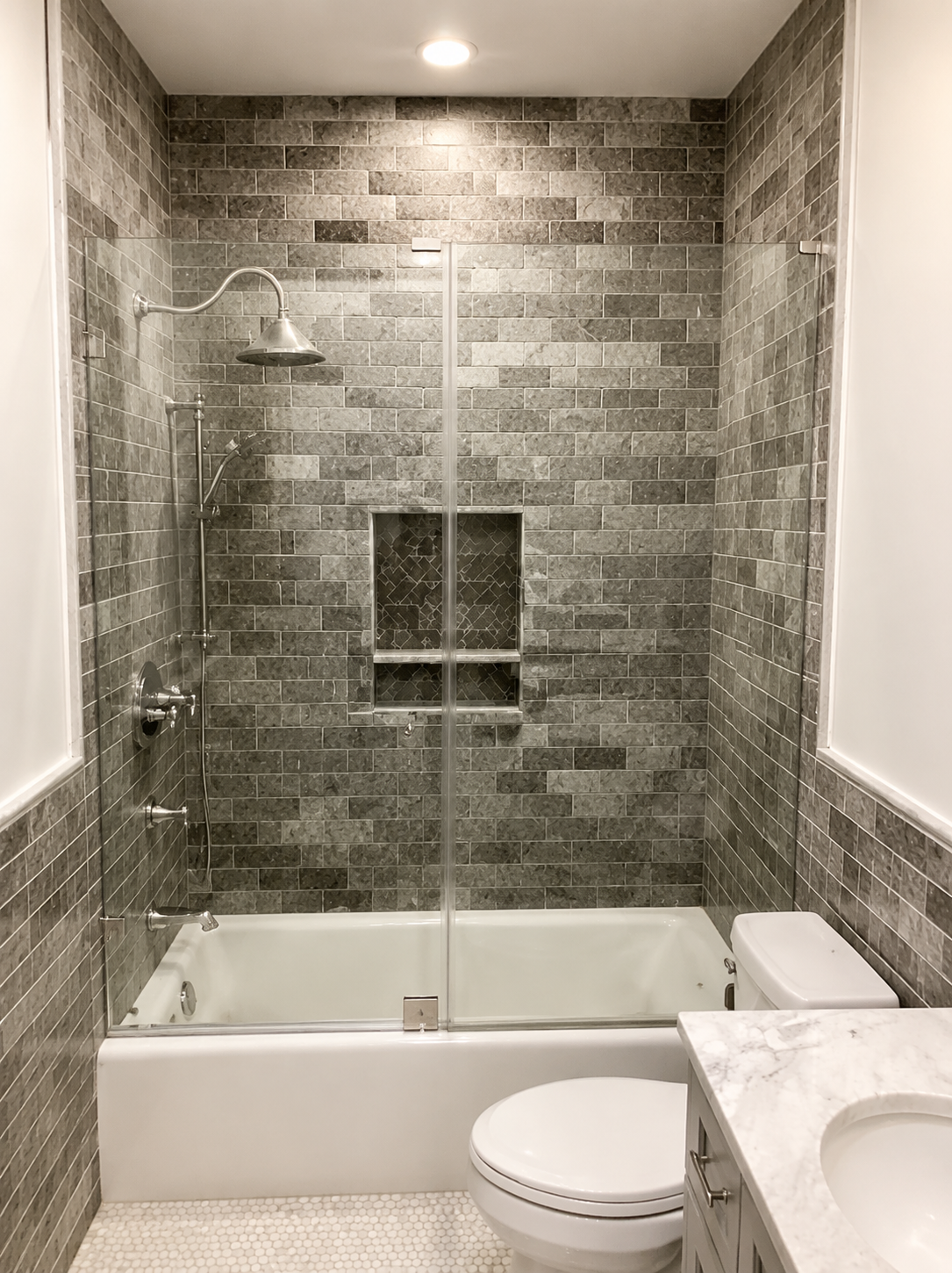 Modern bathroom with a glass shower, gray tile walls, and a white tub and toilet