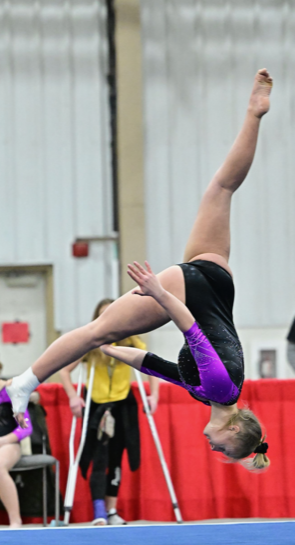 Gymnast mid-air on balance beam, wearing a black and purple leotard, in an indoor gym.