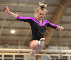 Gymnast mid-air on a balance beam, wearing a black and purple leotard in an indoor gym.