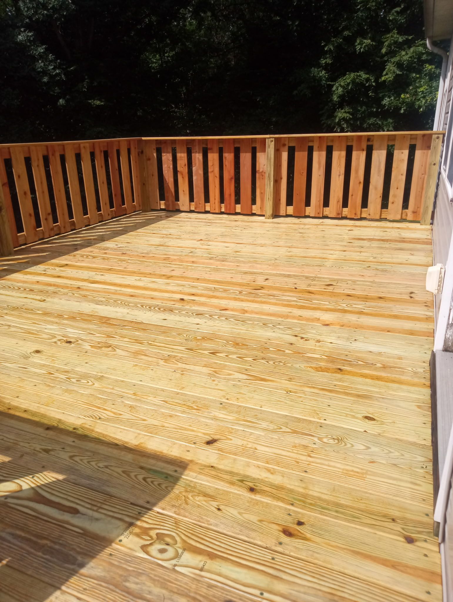 Wooden deck with vertical railing. Sunlit, surrounded by trees.