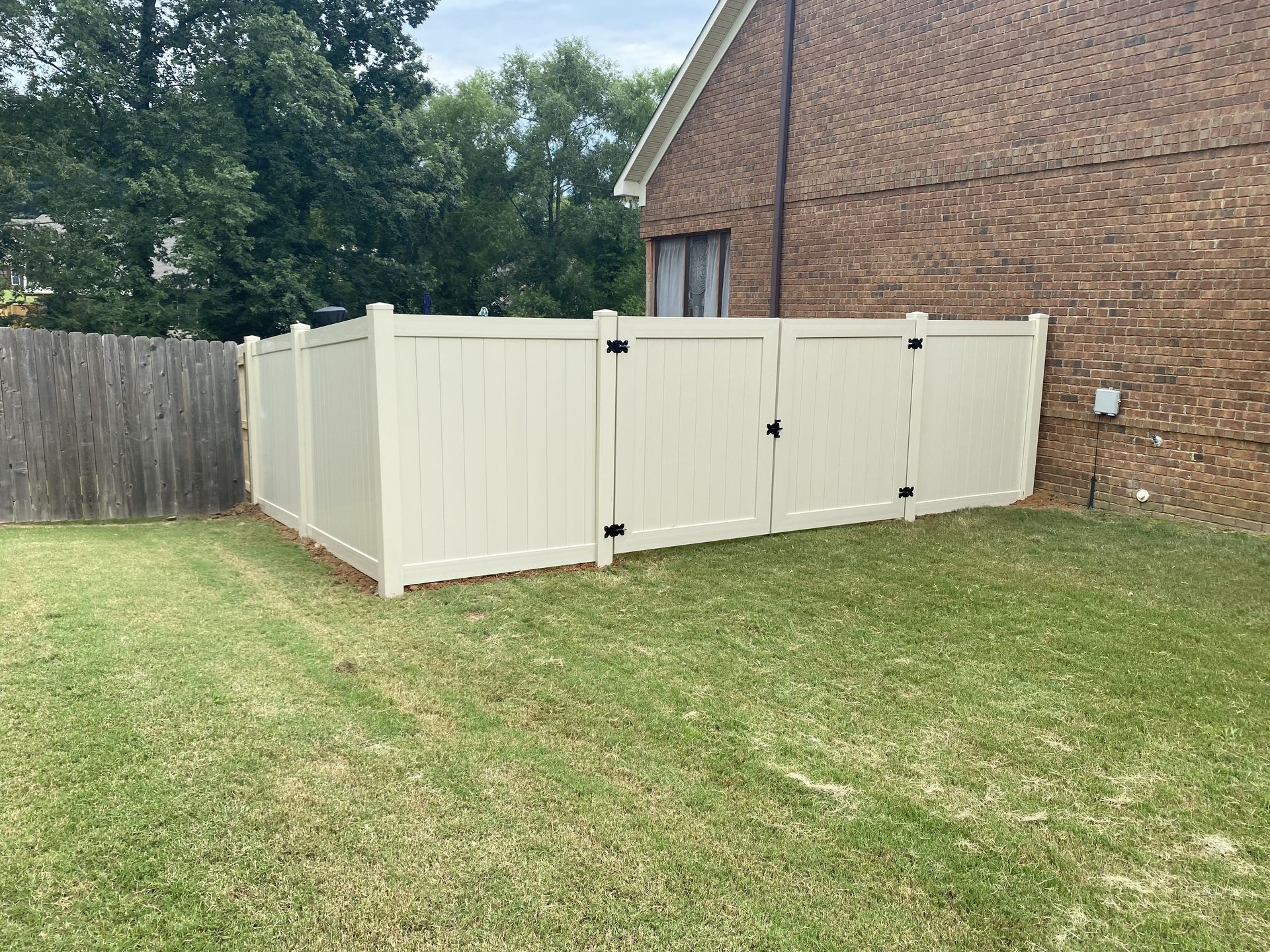 Cream-colored vinyl fence with two gates next to a brick house and wooden fence on a green lawn.