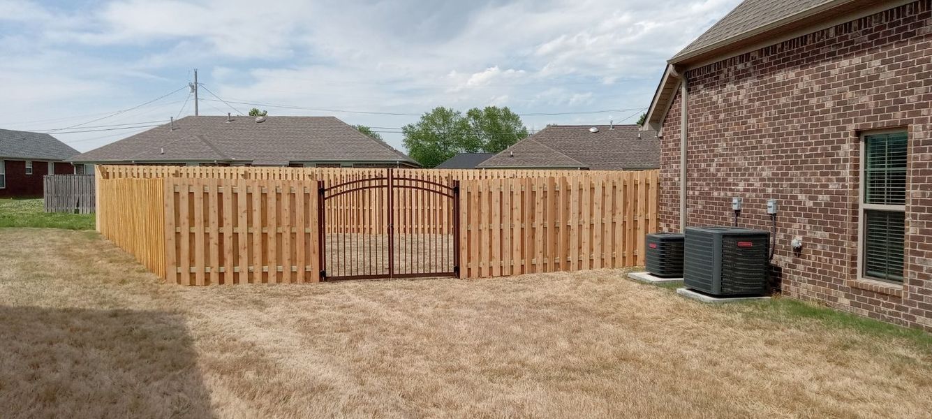 Wooden fence encloses a backyard, with a brick house to the right. Air conditioning units sit beside the house.