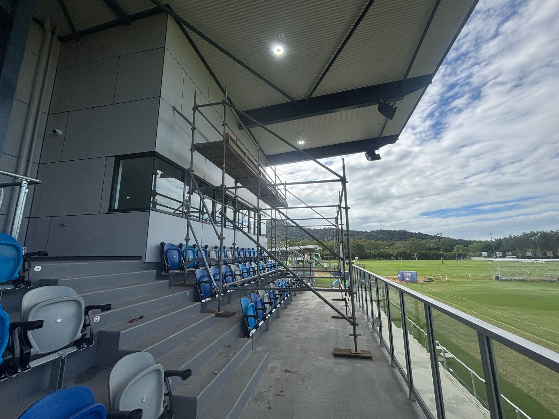 Covered stadium seating beside a soccer field, with blue seats and scaffolding under a roof — Underdog Edge Protection in Redlynch, QLD