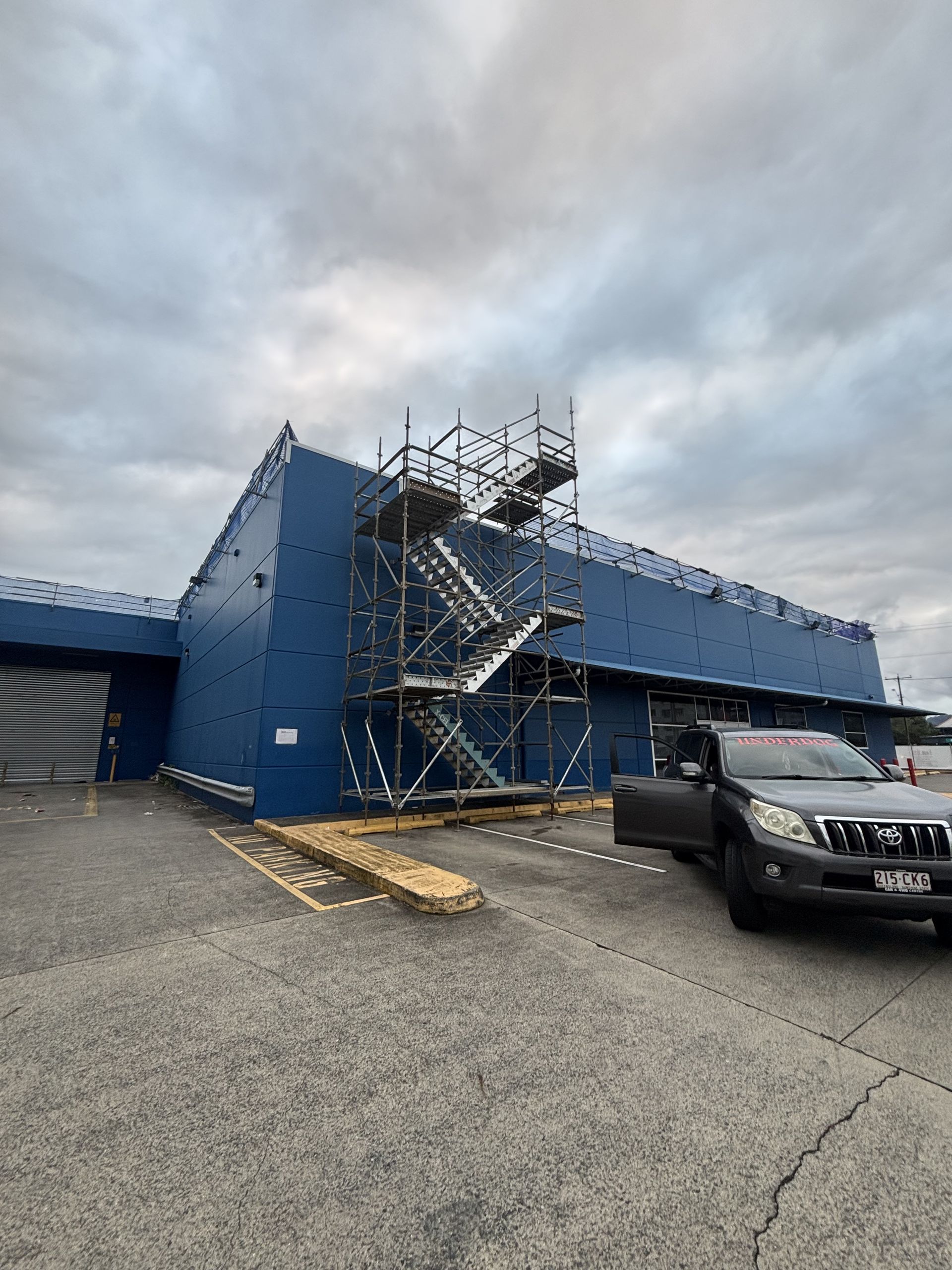 Blue industrial building with scaffolding and a black pickup truck in a gravel lot under cloudy skies — Underdog Edge Protection in Redlynch, QLD