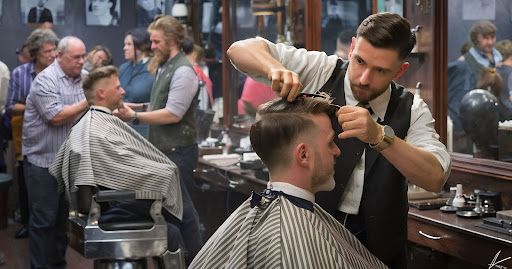 Barber cutting hair of a client at a barbershop. Other barbers and customers are also present.