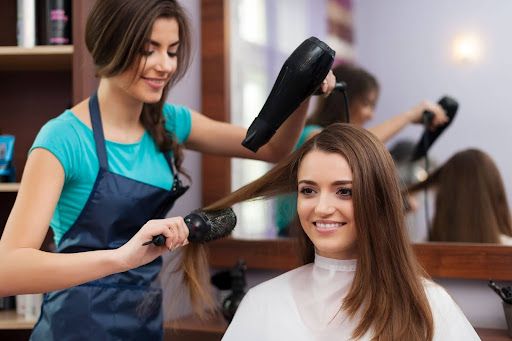 Hairdresser blow-drying a customer's hair in a salon; both smiling, mirror reflection visible.
