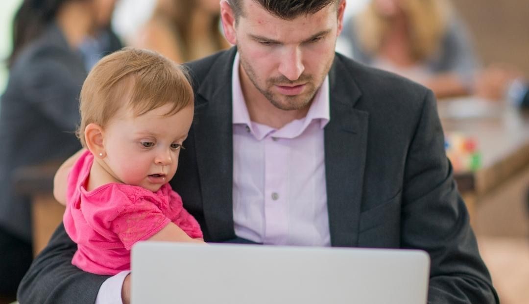 A man is holding a baby while using a laptop computer.