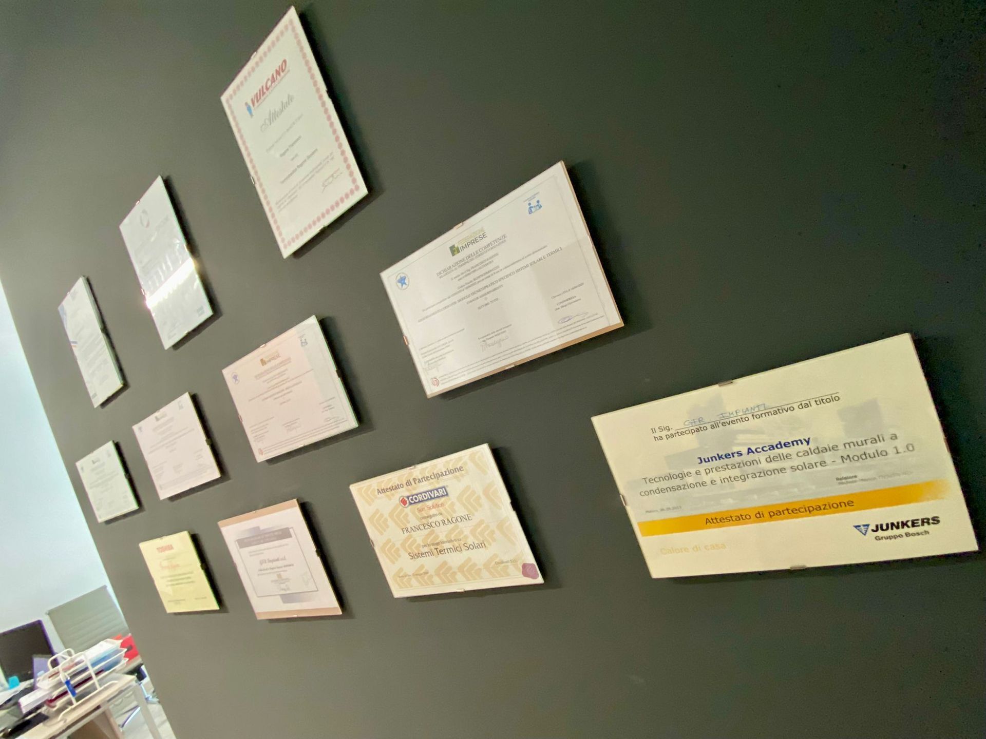 Certificati di merito e riconoscimento esposti su una parete verde scuro in un ufficio.