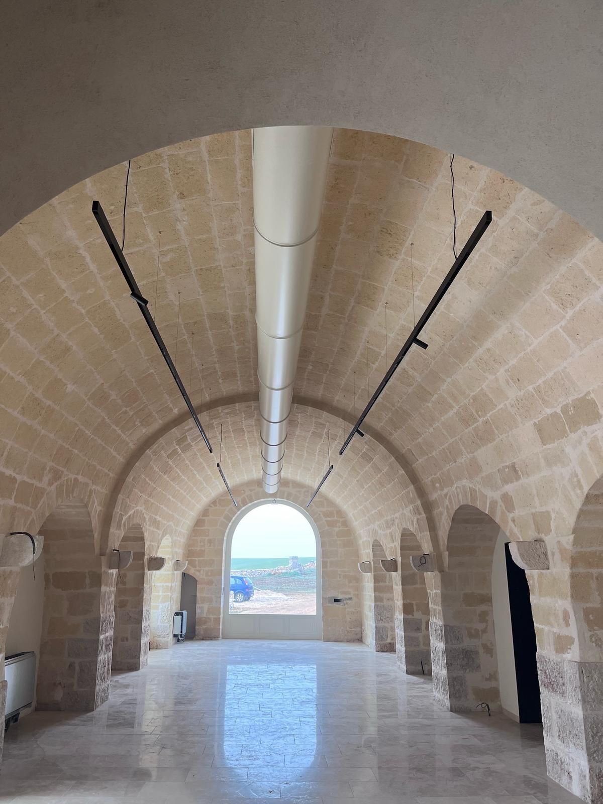 Corridoio ad arco con pareti in pietra, con luce naturale intensa in fondo e lunghe luci appese al soffitto.