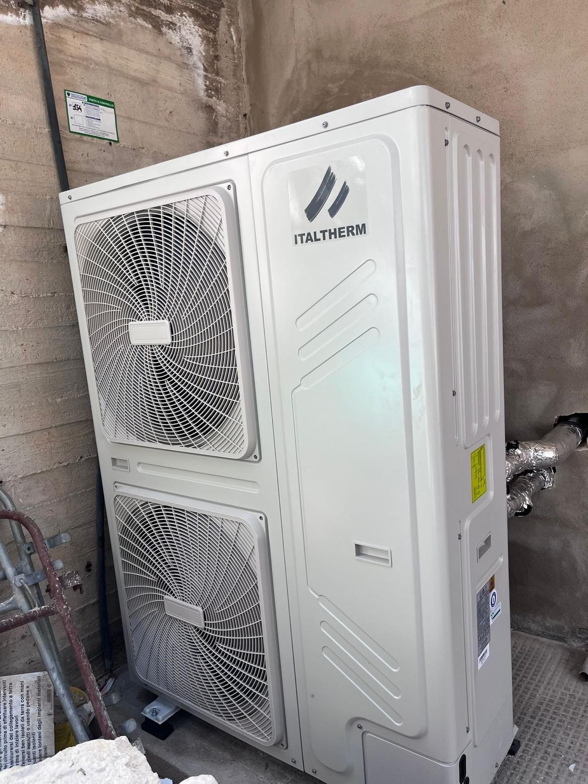 Unità di pompa di calore esterna bianca contro una parete, con due griglie di ventilazione visibili.