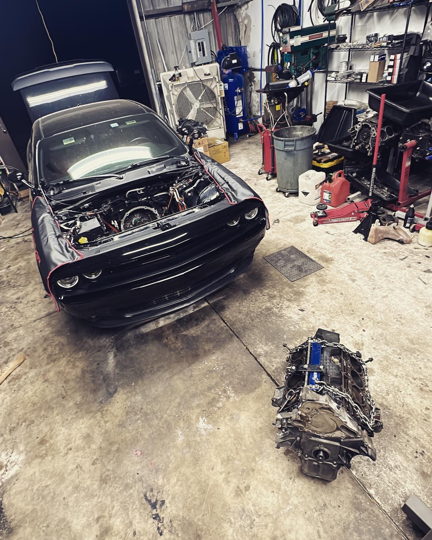 Black Dodge Challenger with the hood open, next to a detached engine in a garage. | YellowHammer Racing