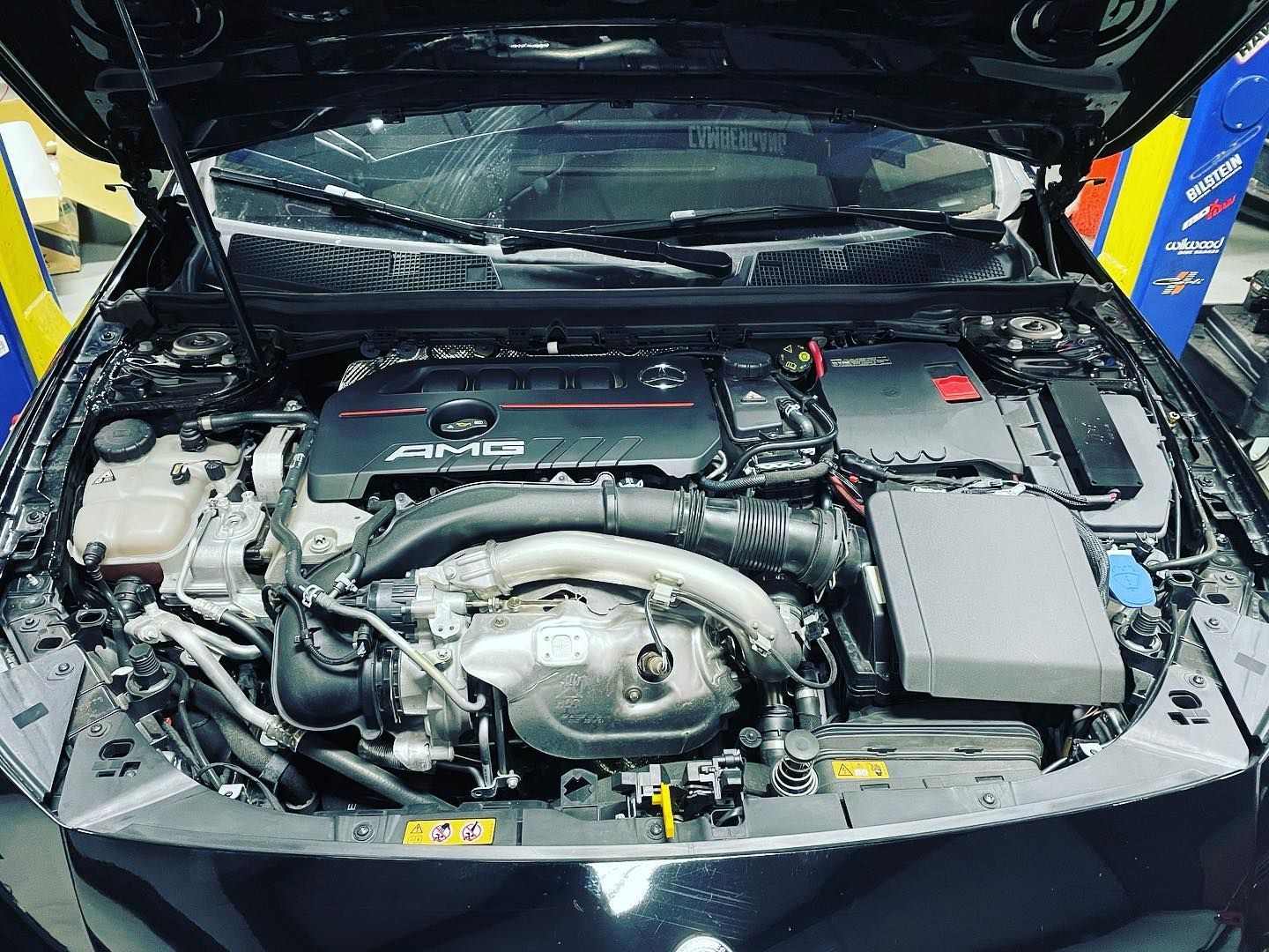 Engine bay of a black car with the hood open. Features the AMG logo and various engine components. | YellowHammer Racing