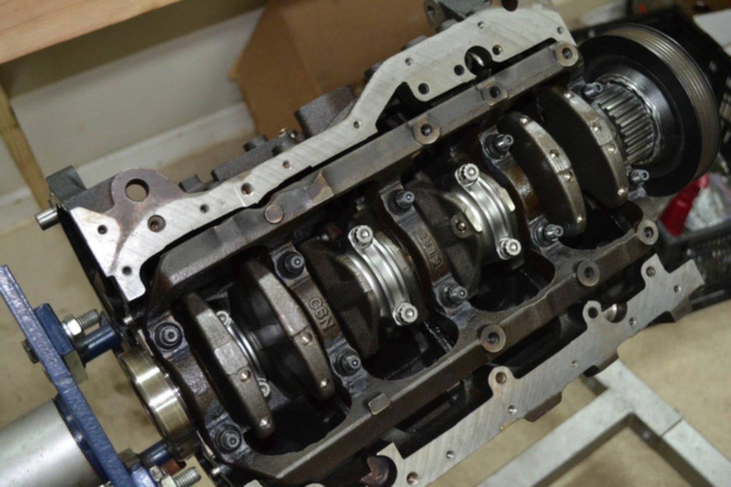 Engine block partially disassembled, showing crankshaft, connecting rods, and bearing caps. Dark metal, industrial setting | YellowHammer Racing