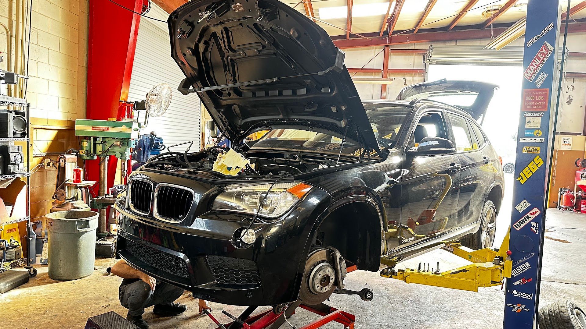 Black BMW SUV with hood open on a lift in a garage. Mechanic working on front wheel.  | YellowHammer Racing