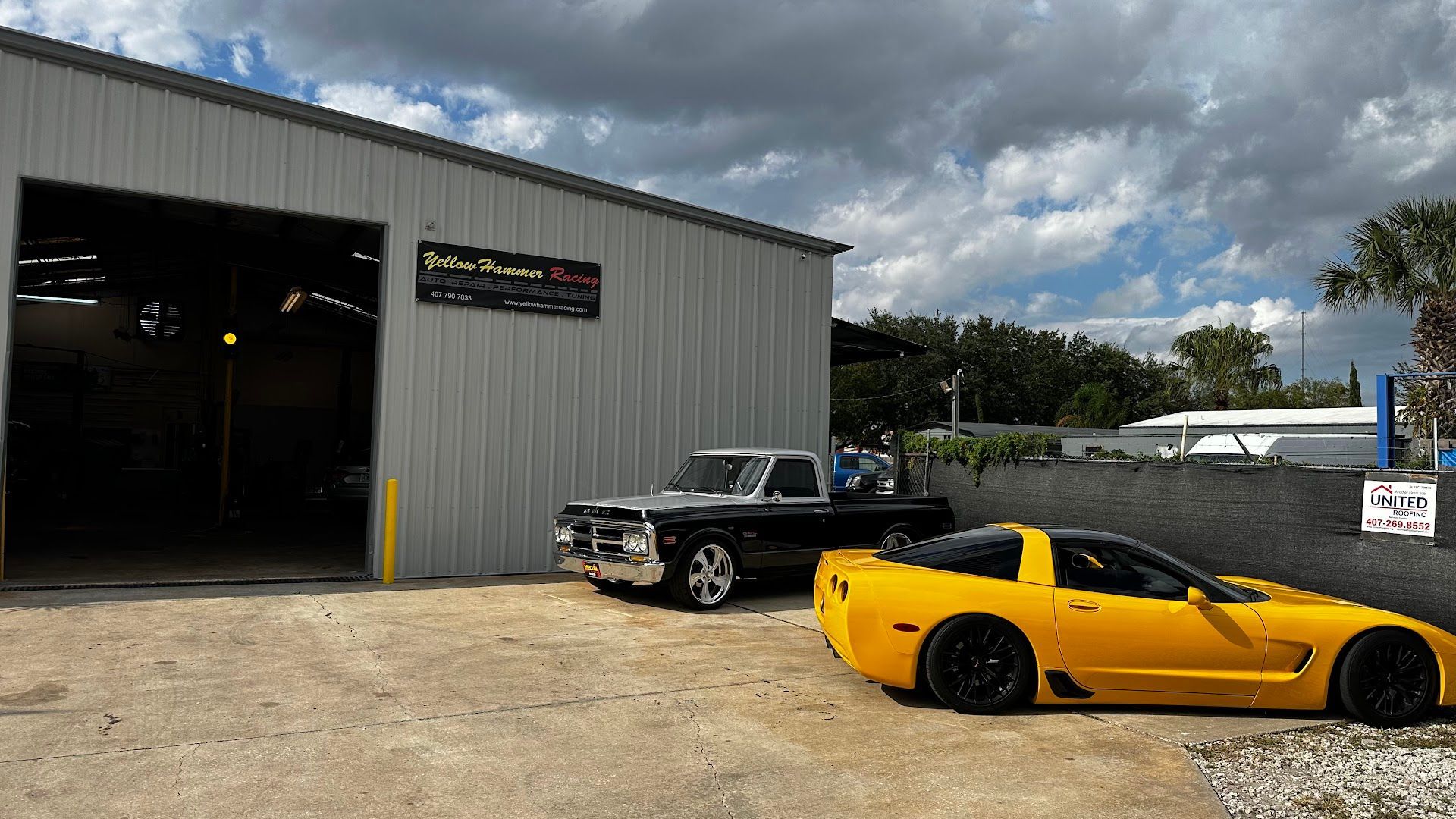 A yellow sports car and black pickup truck parked outside an auto repair shop. Cloudy sky overhead.  | YellowHammer Racing