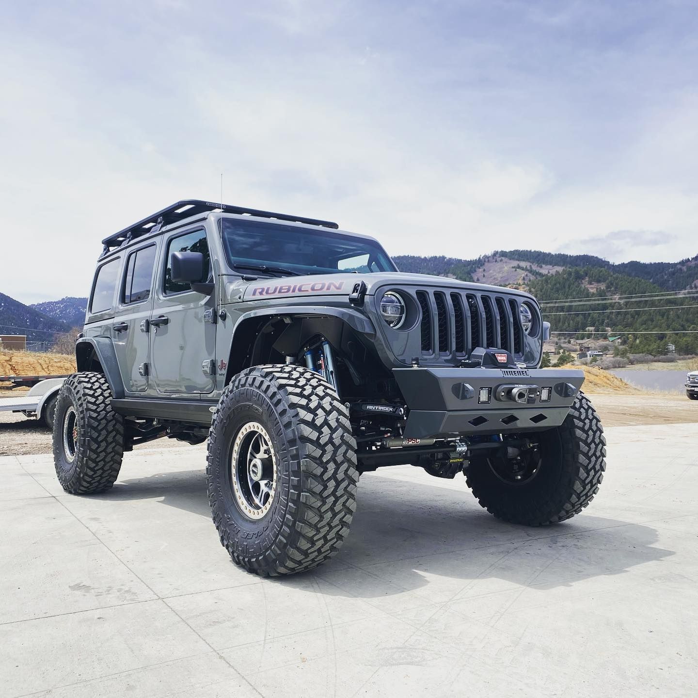 Gray Jeep Wrangler Rubicon with large off-road tires and roof rack, parked on pavement with mountain background. | YellowHammer Racing