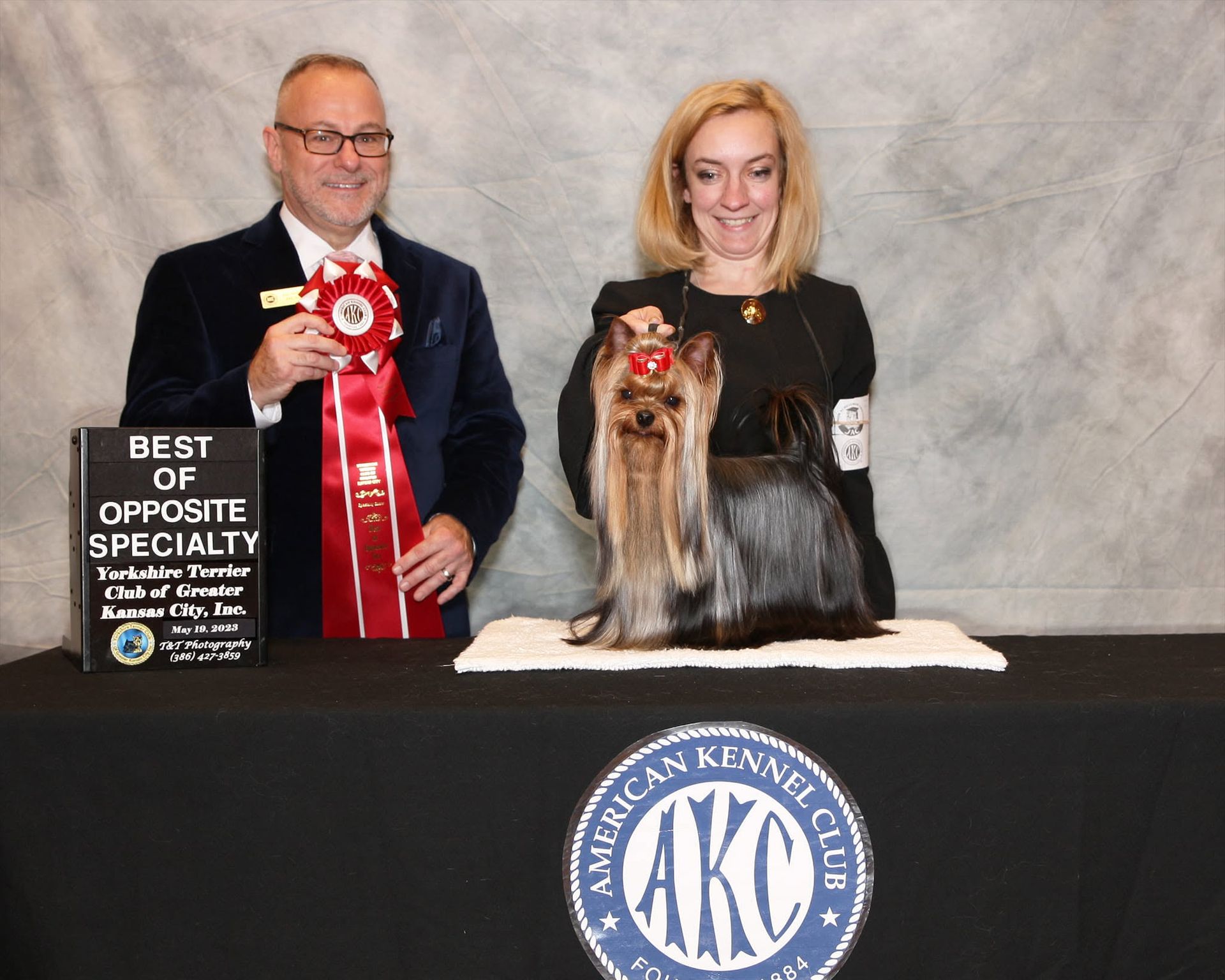 Two people with a long-haired dog at an AKC event, holding a Best of Opposite Specialty award plaque.
