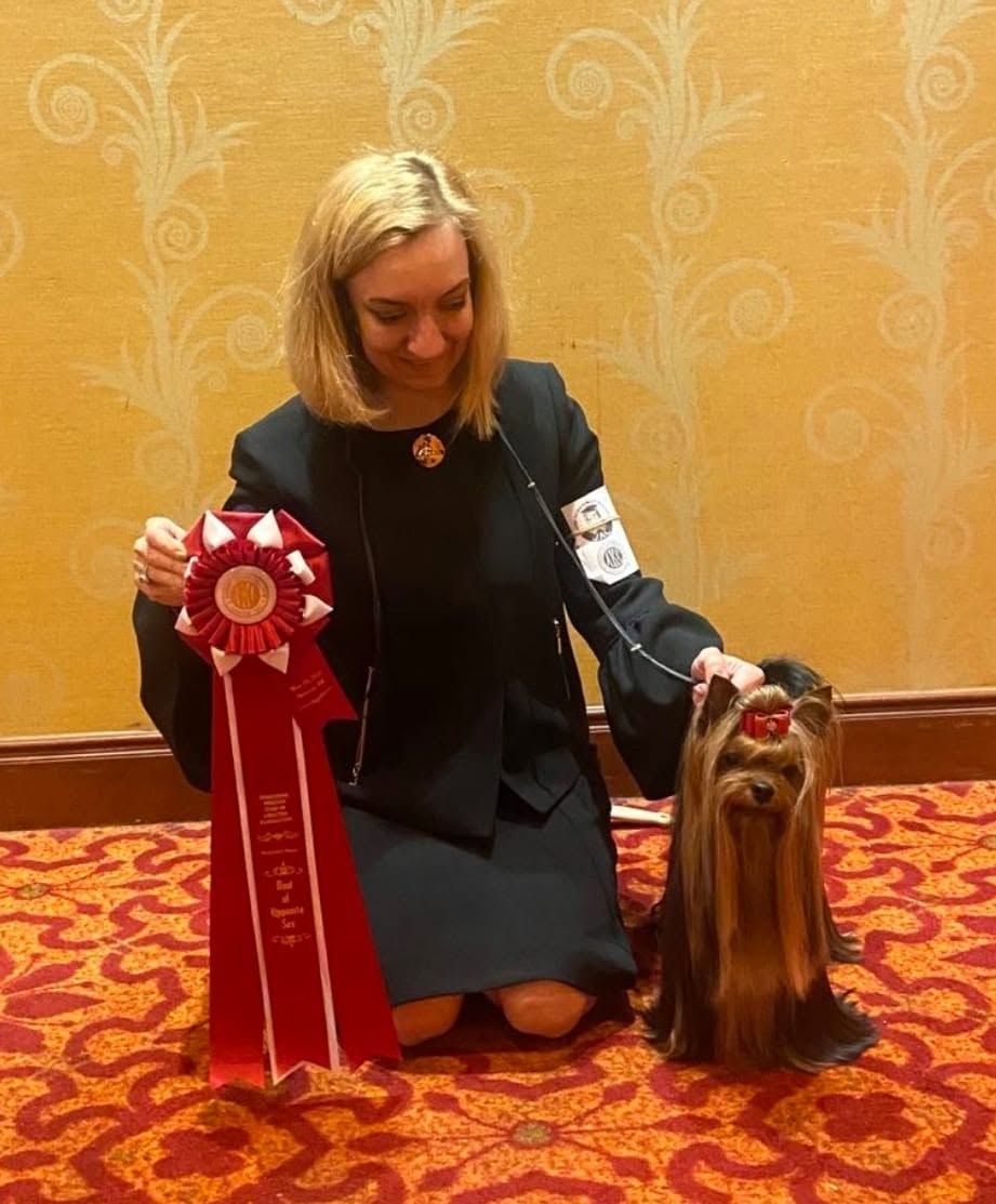 Woman kneels with a prize ribbon beside a long-haired dog at an indoor dog show.