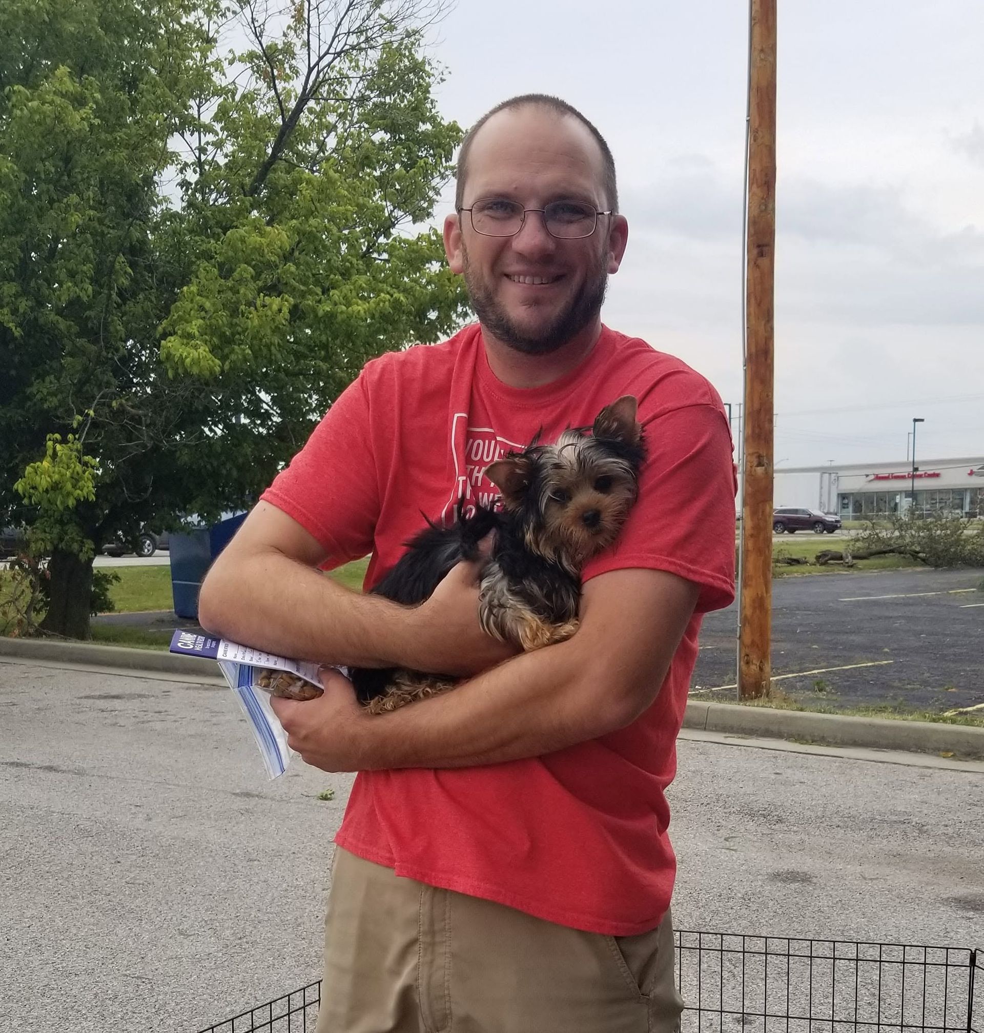 Man in a red shirt holding a small dog outdoors in a parking lot