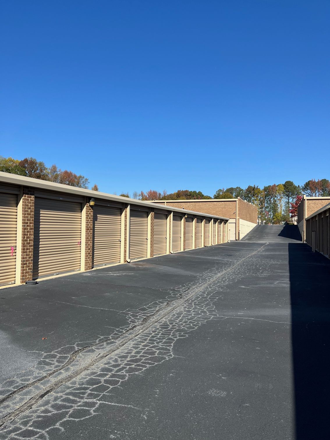Storage units with brown doors line a black asphalt road under a clear blue sky.