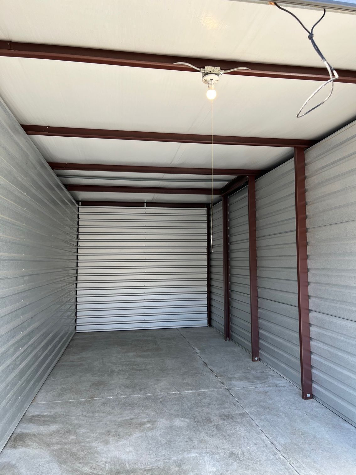 Empty storage unit with a metal door, walls, and brown support beams. Concrete floor, overhead light.