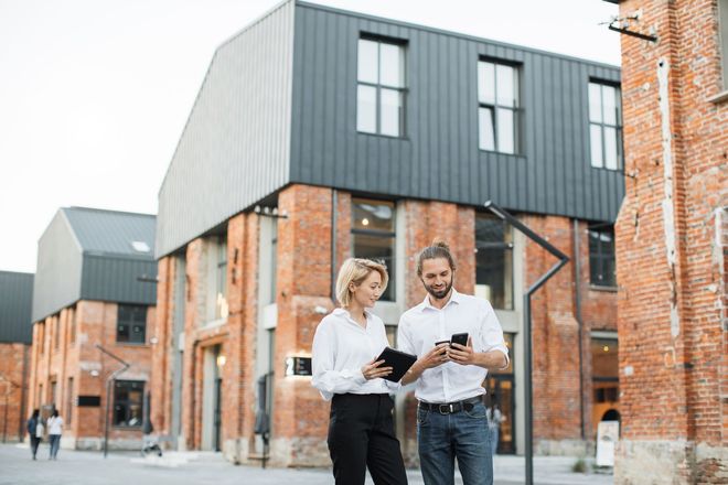 A woman with a tablet and a man with a smartphone discussing work outside a modern brick building.