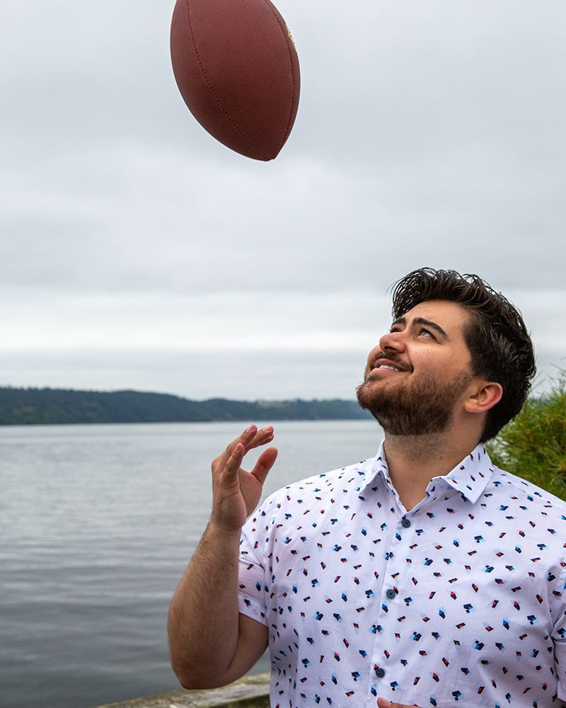 Man throws a football upward near a lake, smiling, wearing a patterned shirt. Cloudy sky.