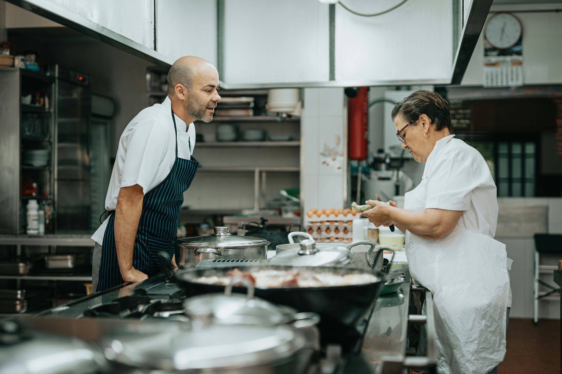 Chef in blue apron looks on as cook adds to food on stovetop in kitchen.