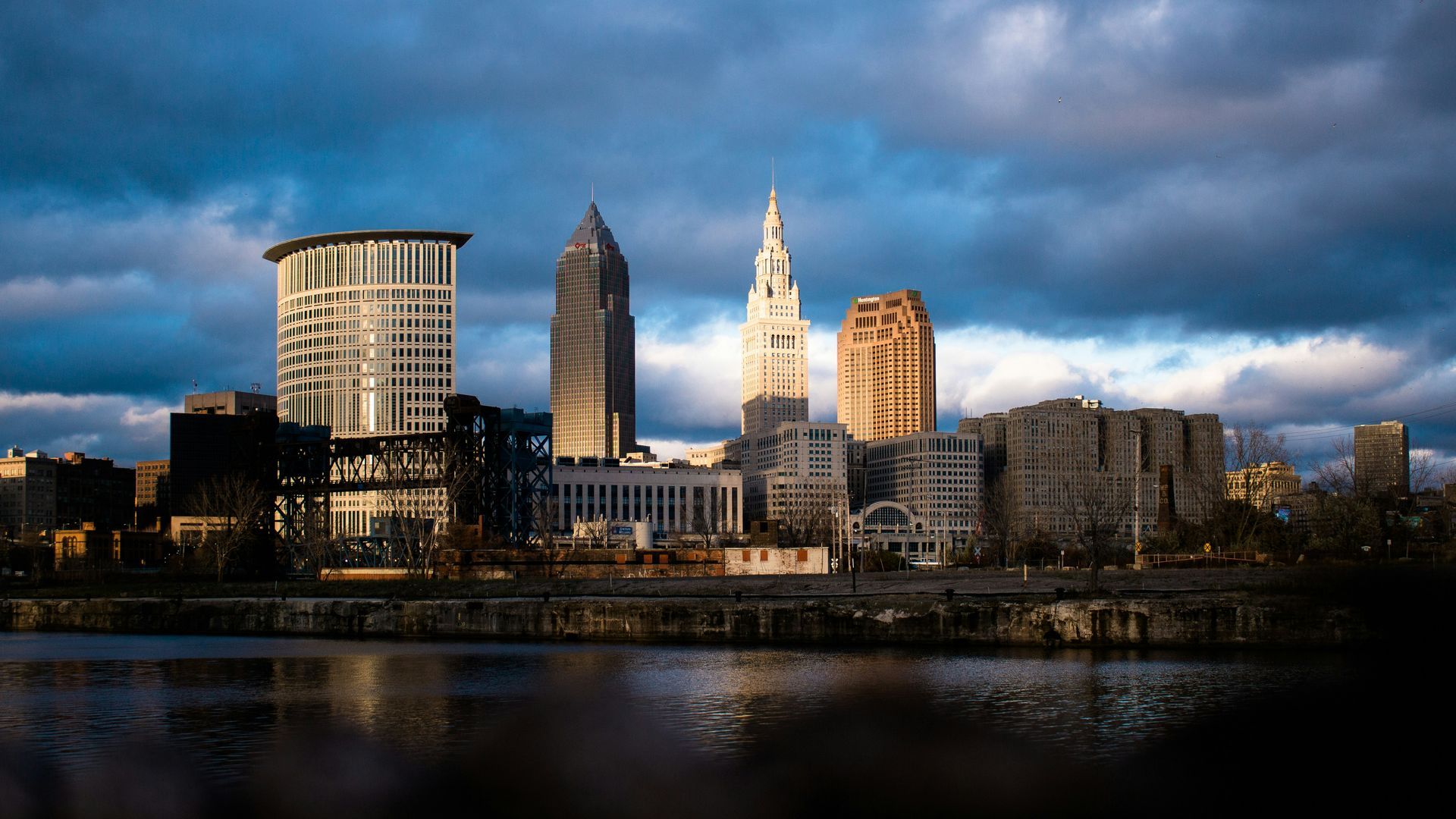 Cleveland skyline at dusk, buildings reflecting in the water, under a cloudy, blue sky.