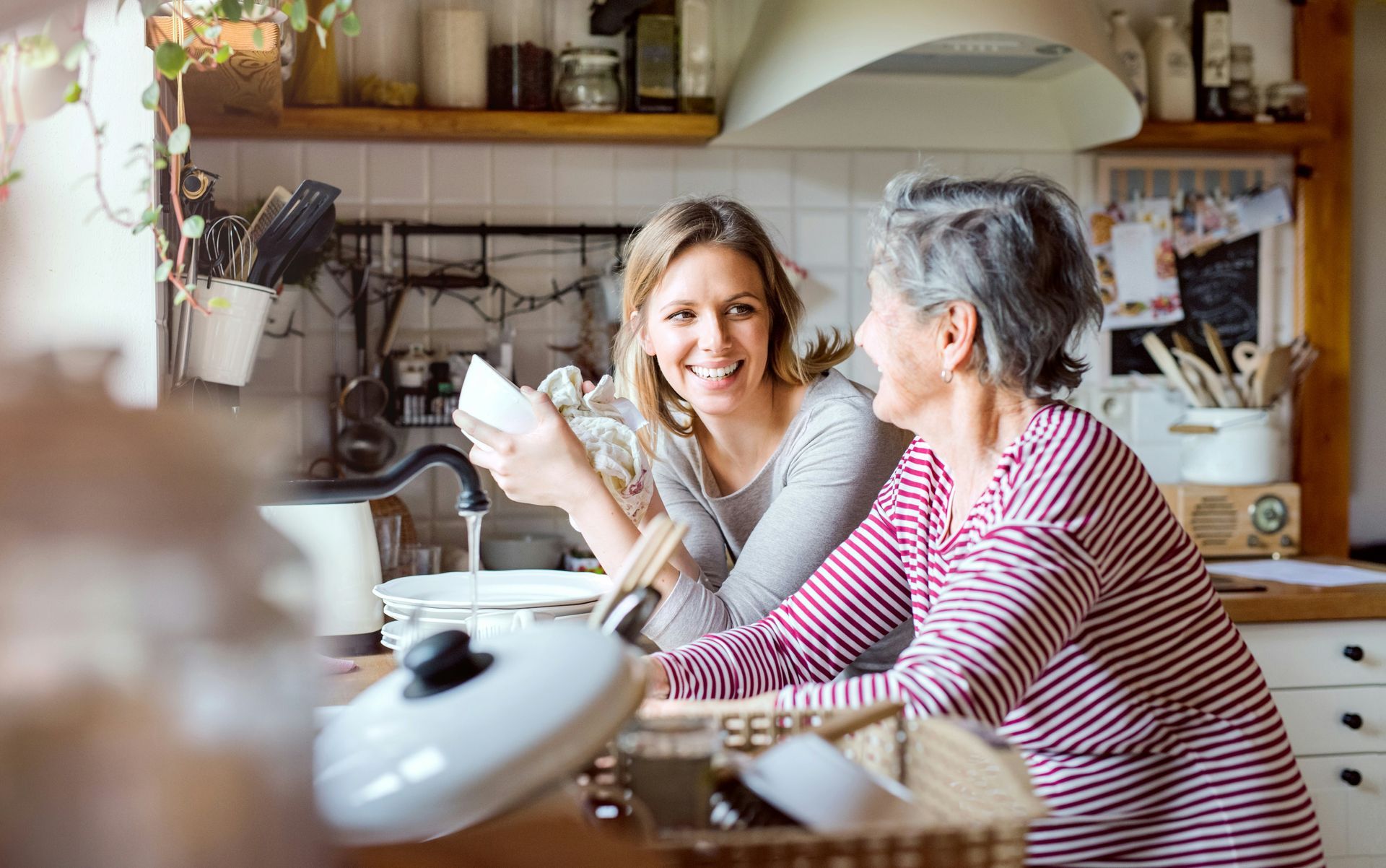 Woman washes dishes while older woman watches in a bright kitchen.