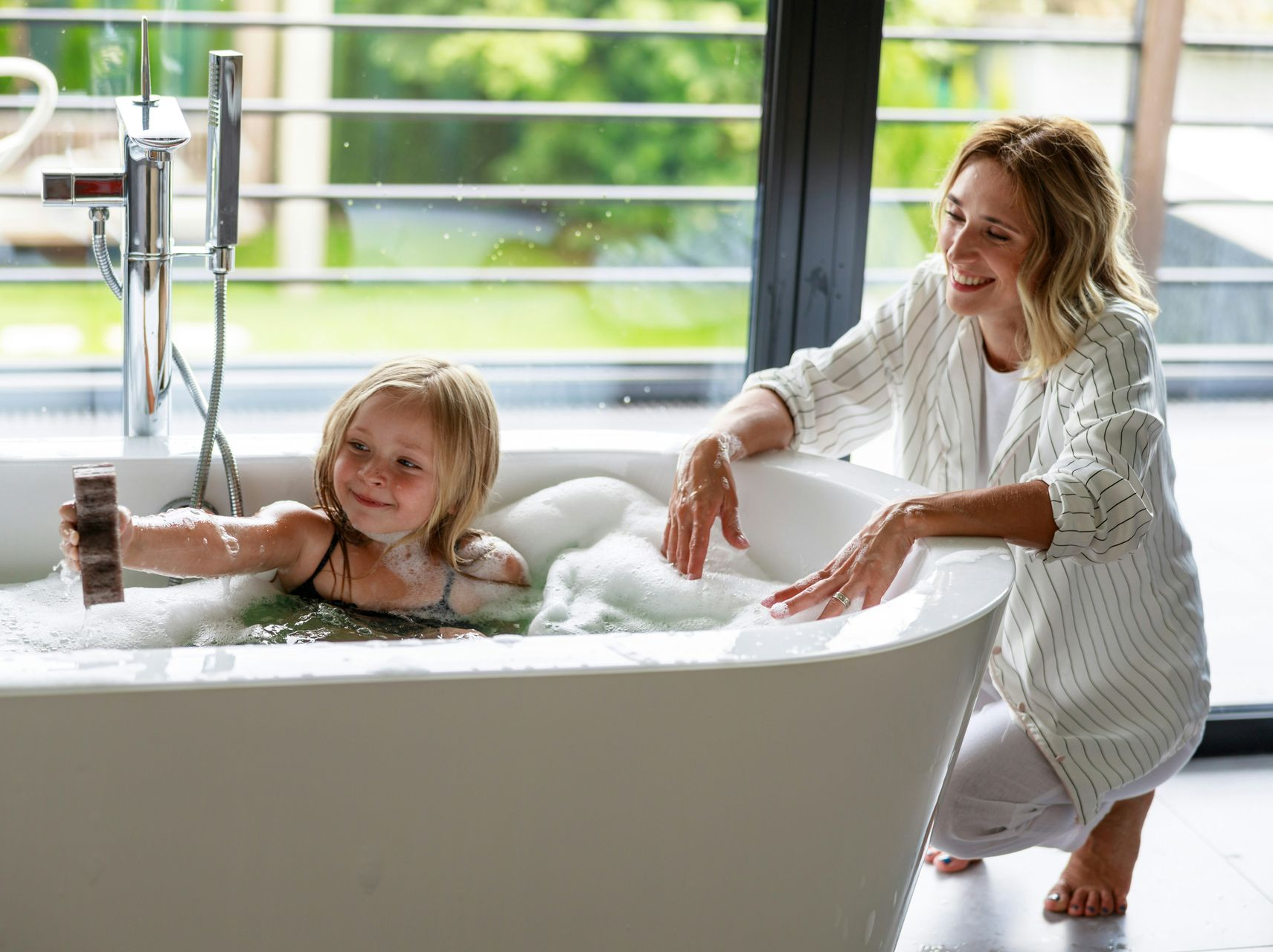 Woman kneels beside a white bathtub, smiling at a child playing in bubbles. Large window in background.