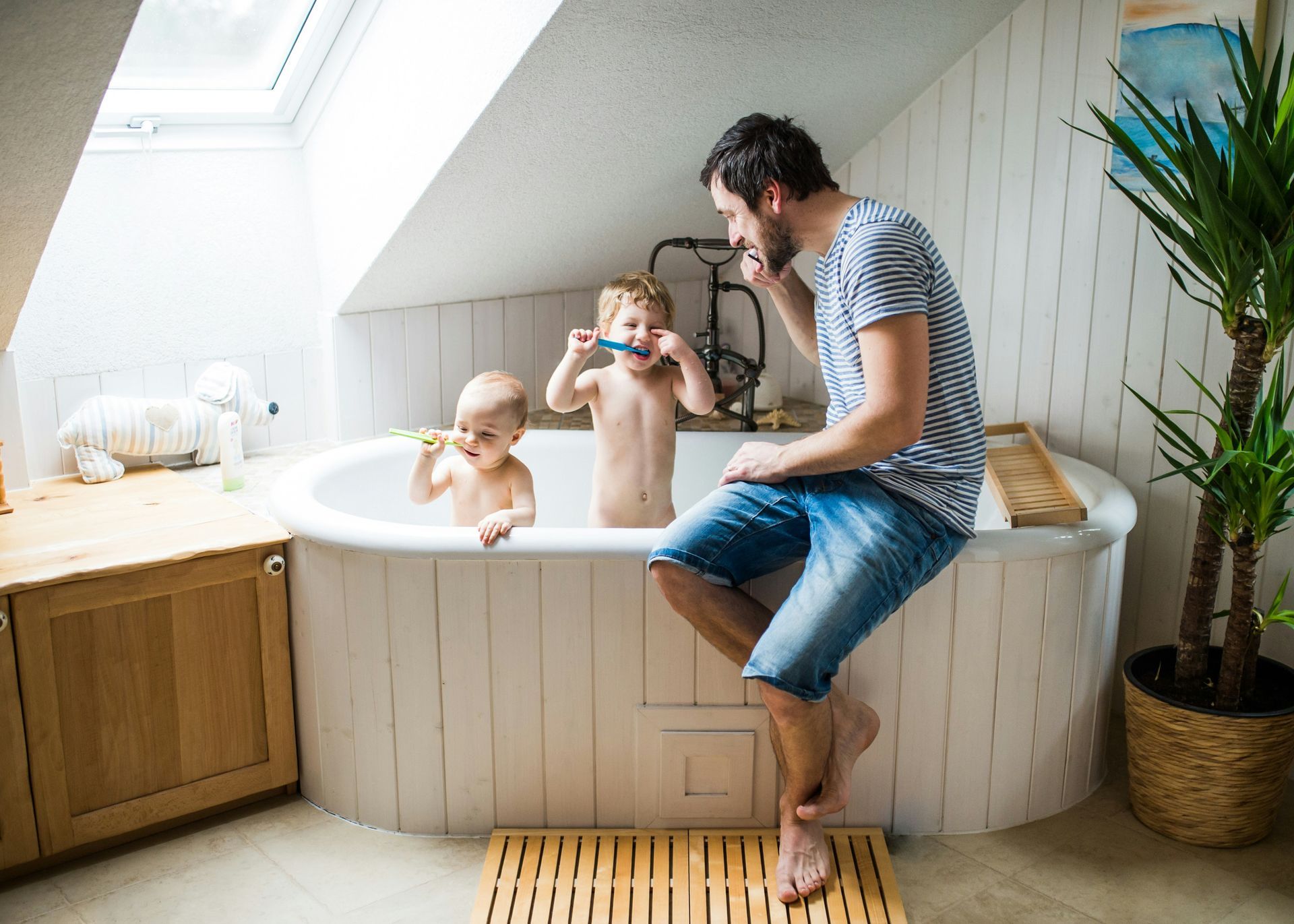 Man brushing teeth while two children play in a bathtub. Bathroom with natural light.