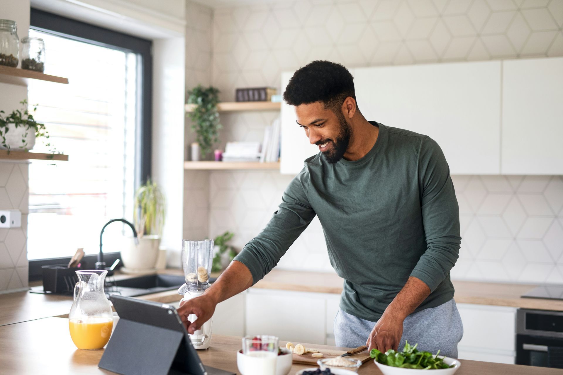 Man in kitchen looking at tablet, preparing food with a blender nearby.