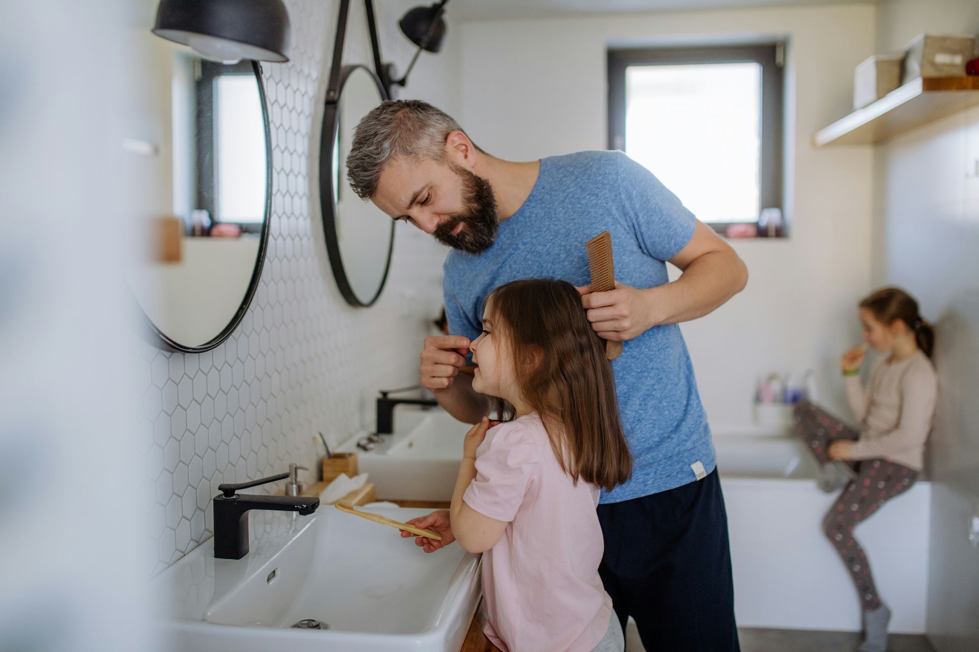Father combing his daughter's hair in a bathroom; a second child in the background.